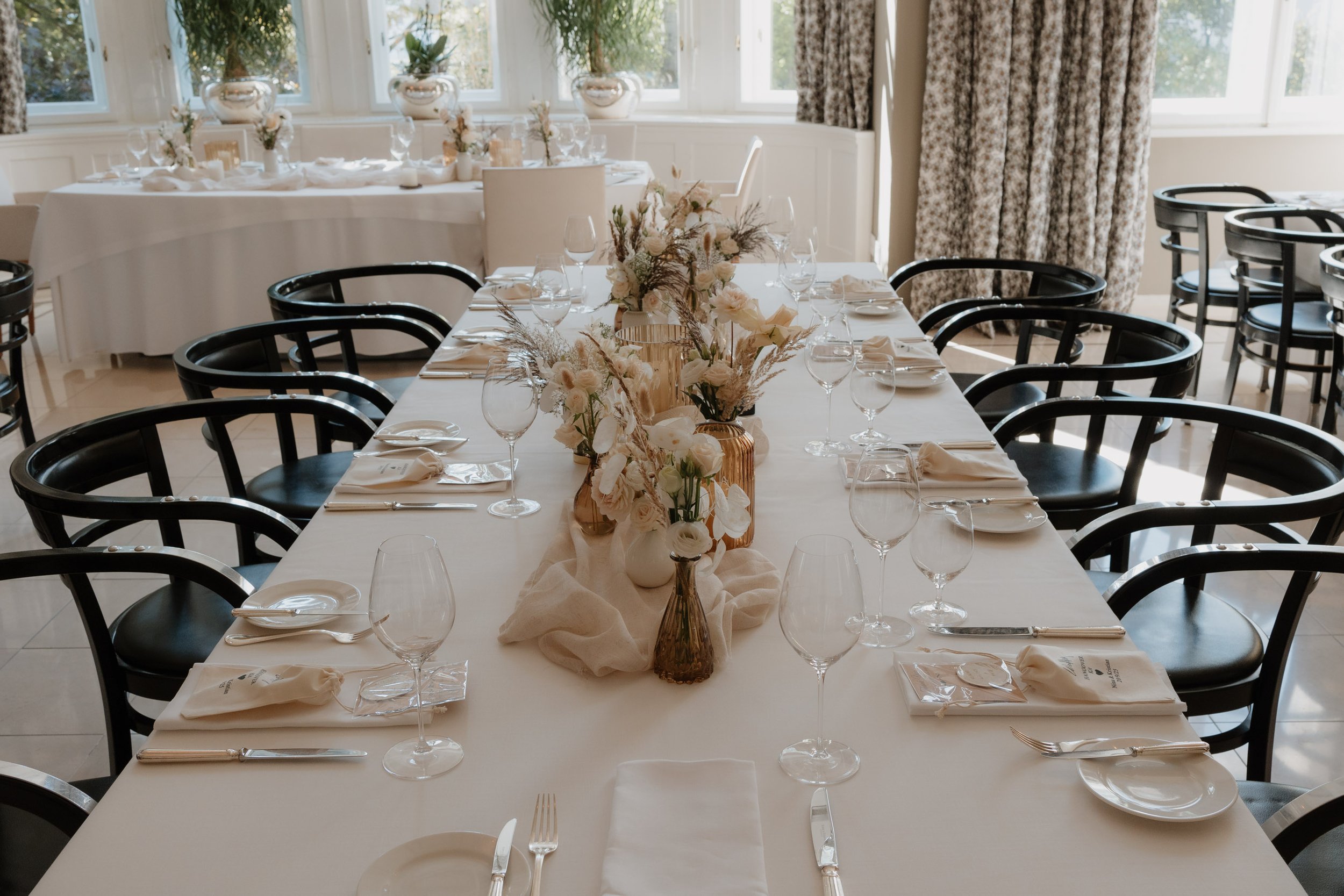 Detailaufnahmen. Ein festlich gedeckter Tisch mit weißen Tischtüchern, Blumenarrangements in Vasen, Gläsern und Besteck in einem hellen Raum mit großen Fenstern und Vorhängen.