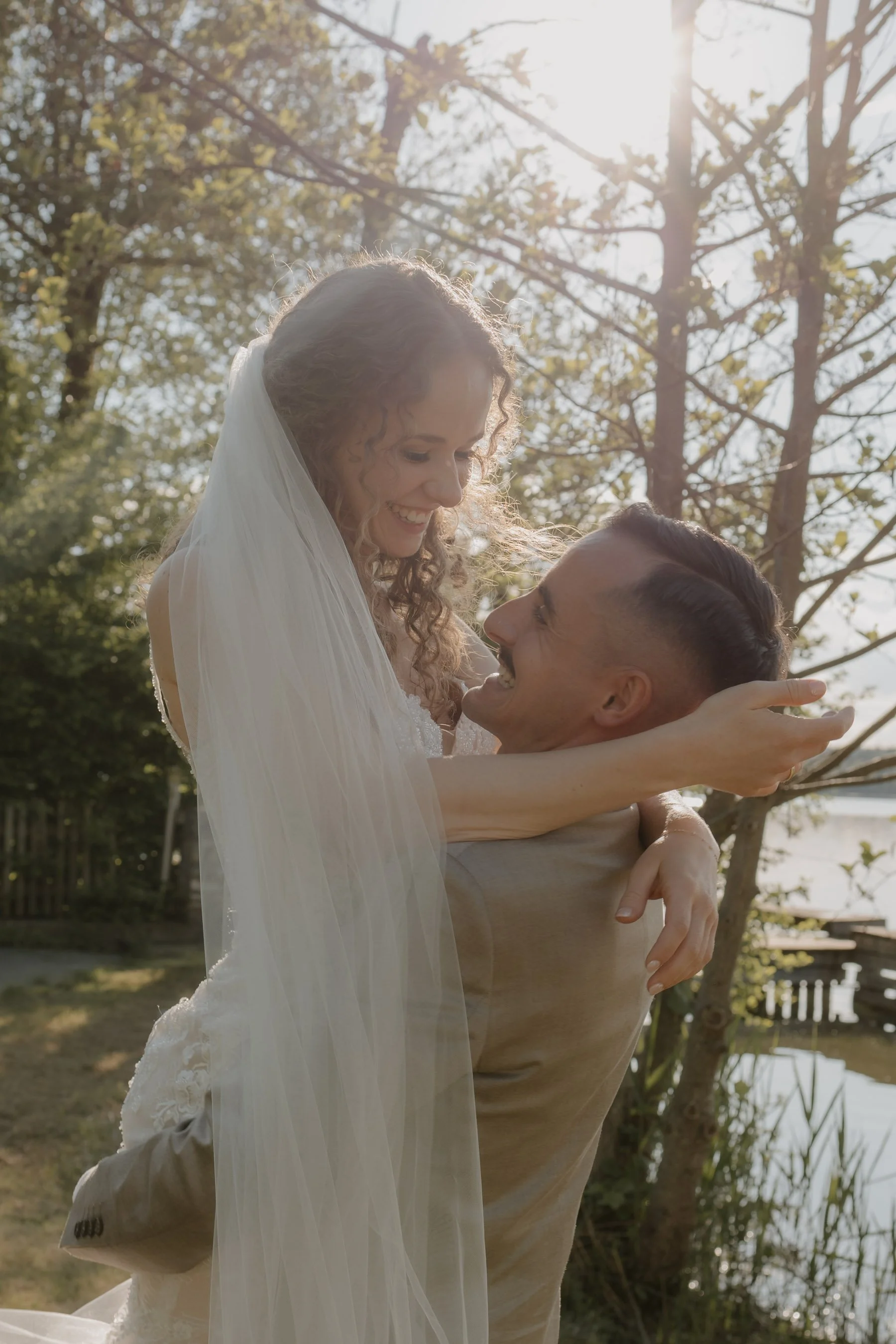 Ein glückliches Paar bei einer Hochzeit im Freien, das sich umarmt und sich gegenseitig ansieht, mit Sonnenschein und Bäumen im Hintergrund.