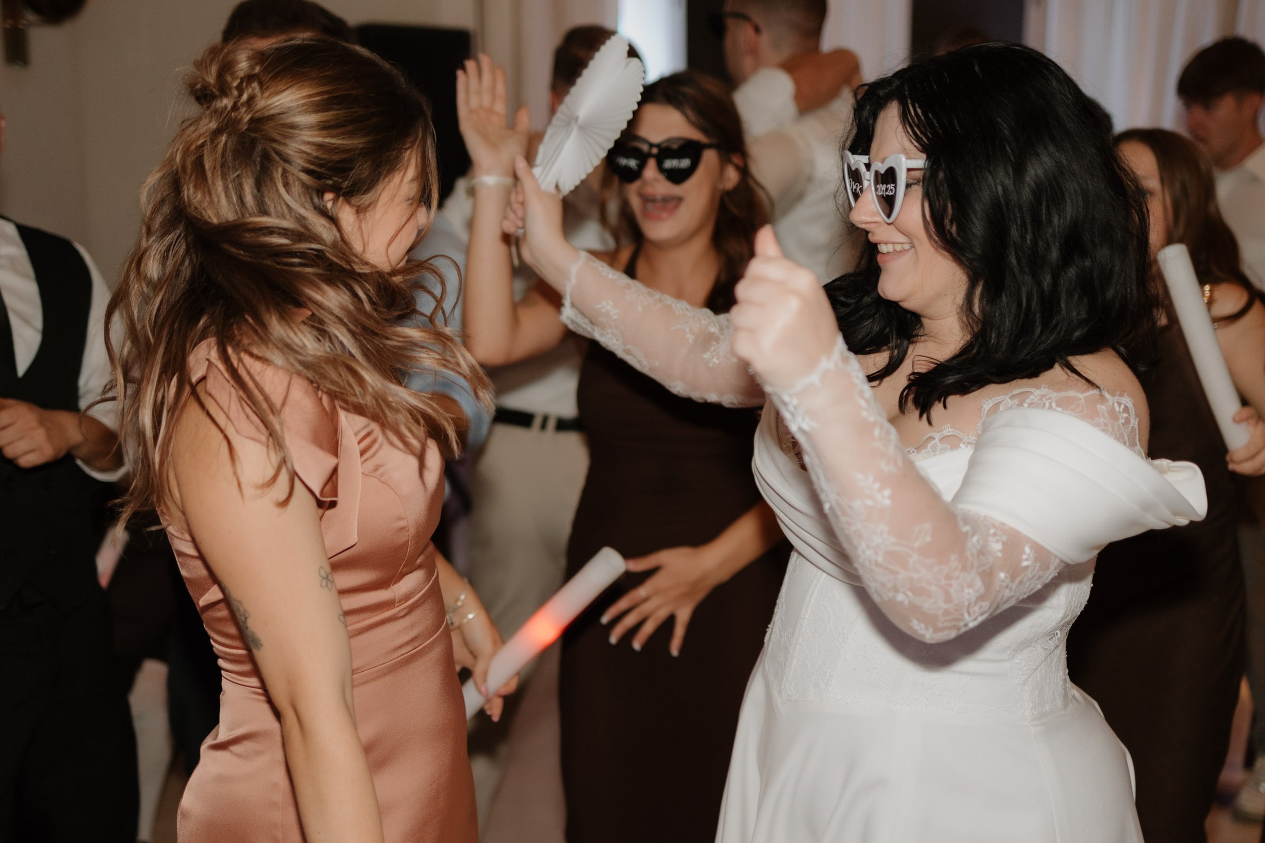Zwei Frauen tanzen auf einer Party. Die Frau auf der linken Seite trägt ein satiniertes rosa Kleid mit kurzen Ärmeln, hat lange, gewellte braune Haare und Tätowierungen am Arm. Die Frau auf der rechten Seite ist in einem weißen Brautkleid, trägt Herz