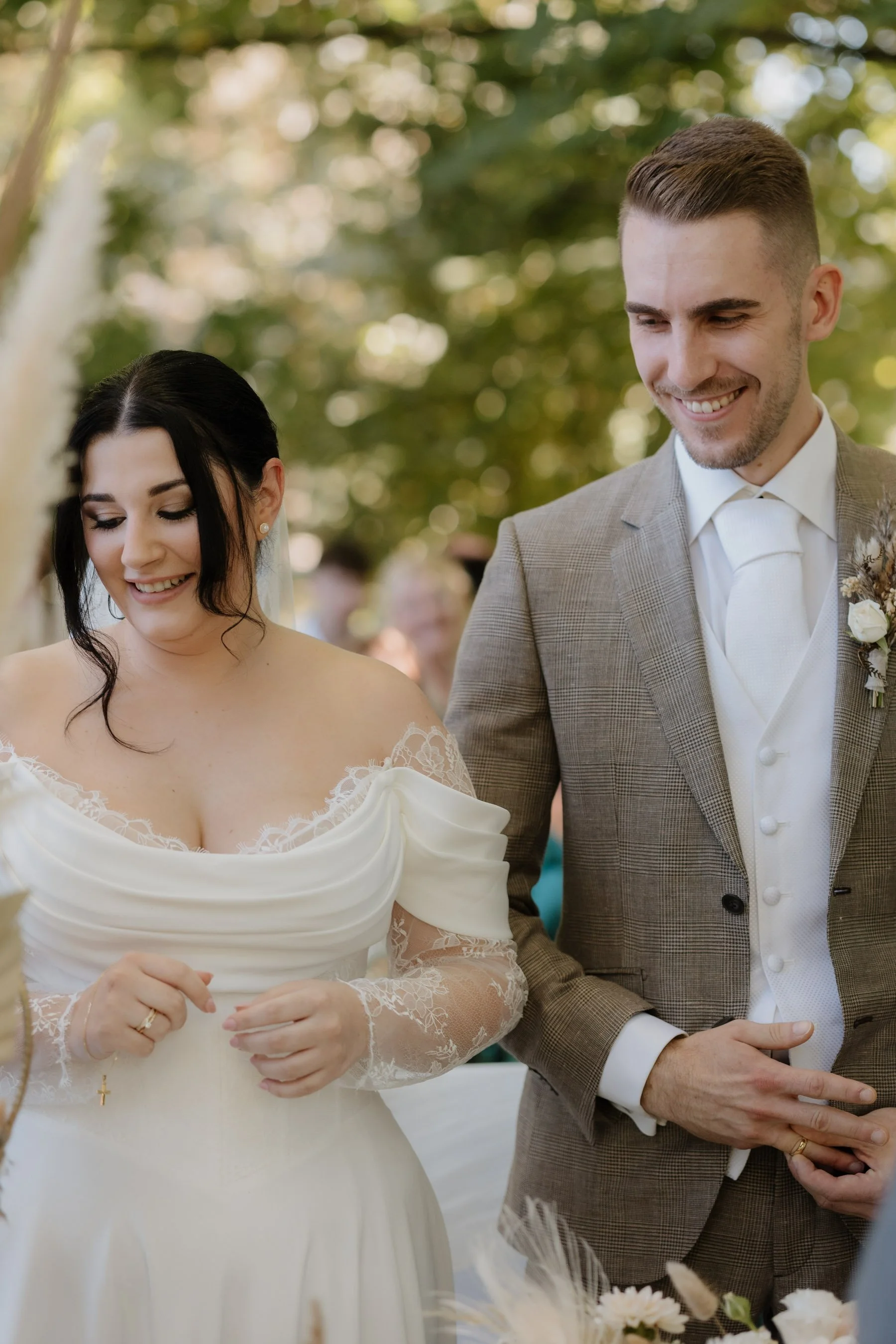 Ein Brautpaar bei einer Hochzeit, die Frau trägt ein weißes Hochzeitskleid mit Lace-Details, der Mann trägt einen grauen Anzug mit Weste und Krawatte, beide lächeln während des Zeremonie.
