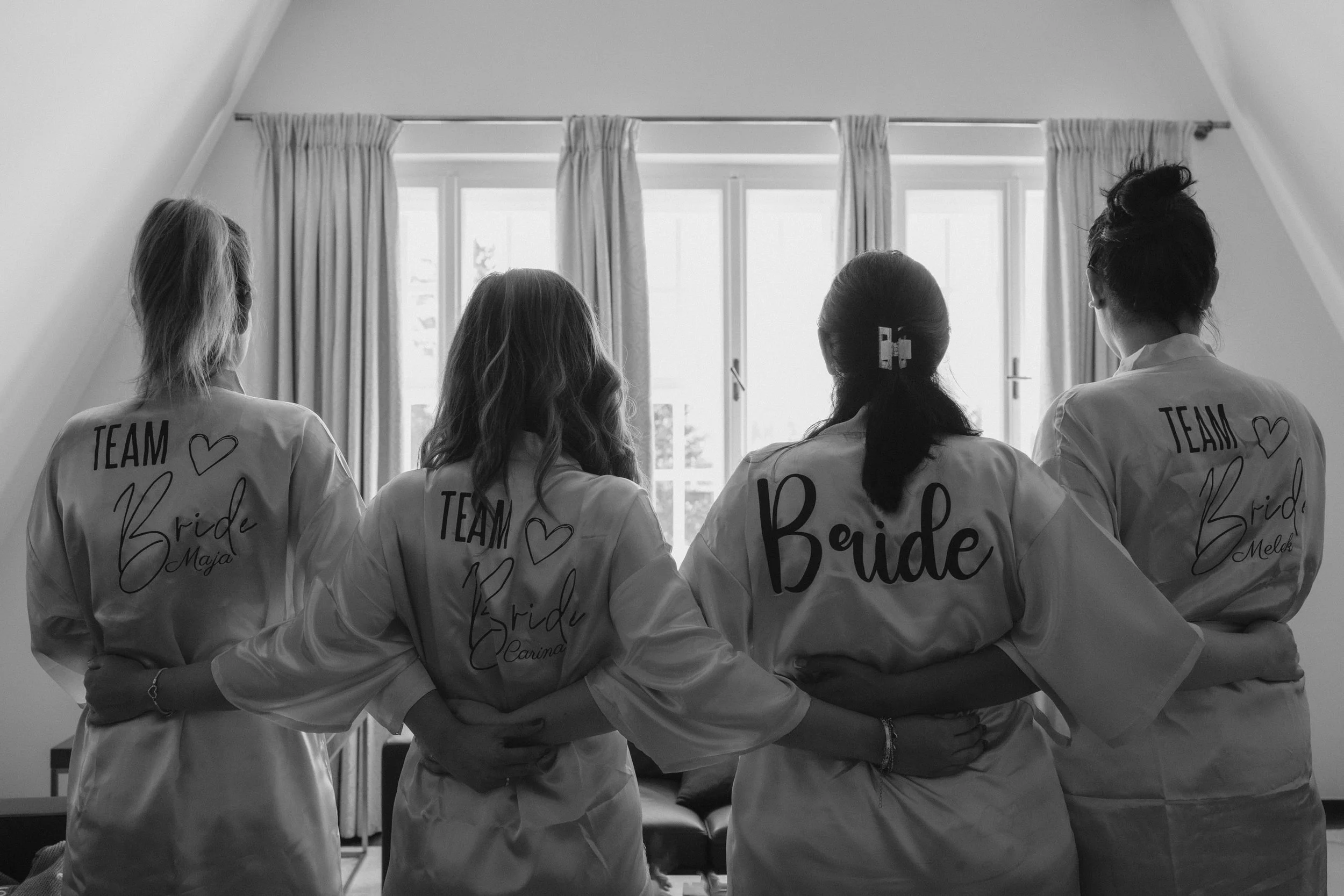 Vier Frauen stehen in einem Raum und schauen durch große Fenster. Sie tragen weiße Jacken mit dem Schriftzug 'TEAM Bride' und Herzen. Es ist eine festliche Stimmung vor einer Hochzeit.