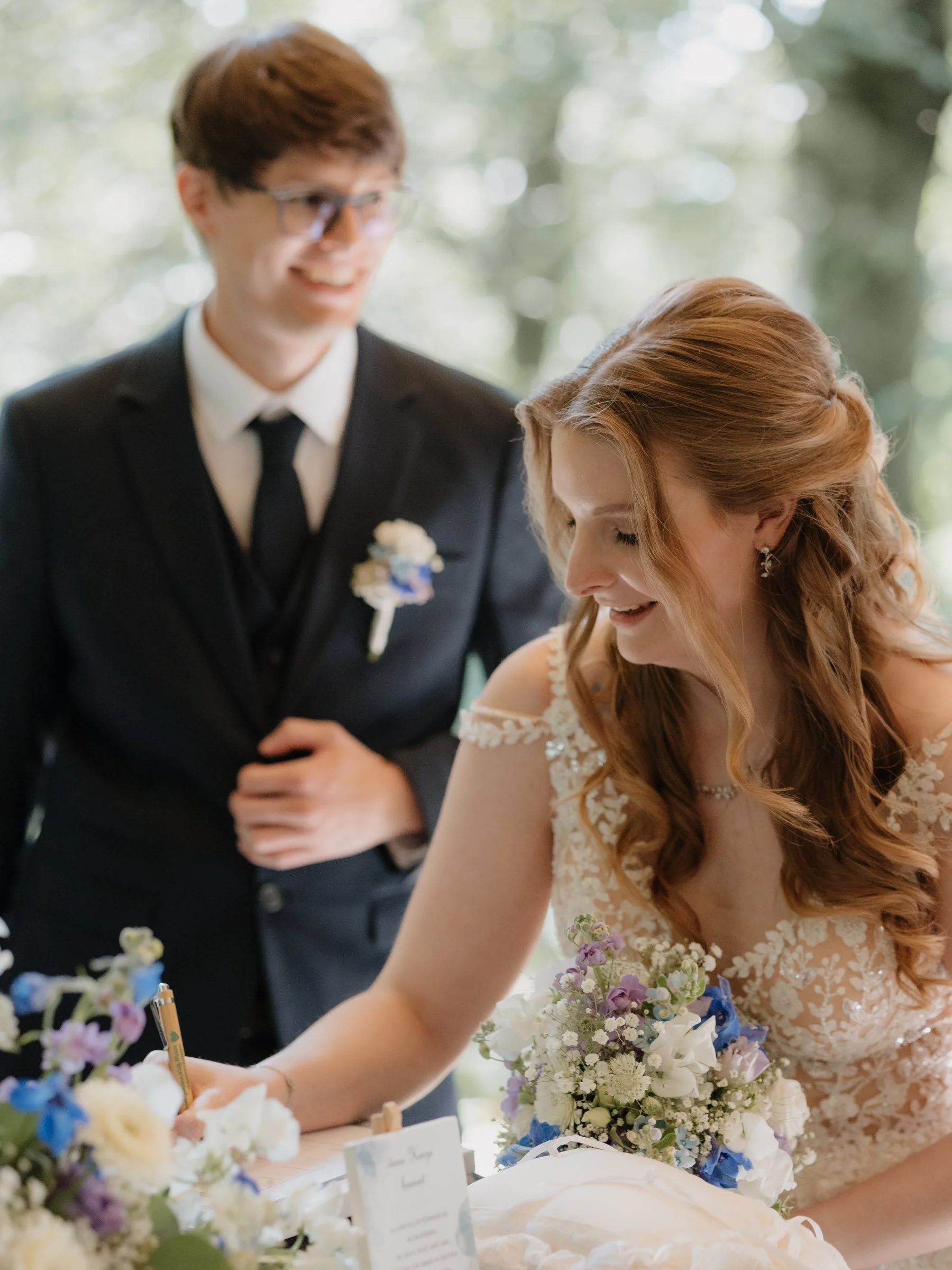 Ein Brautpaar bei der Hochzeitszeremonie, die Braut trägt ein weißes Spitzenkleid und hat Locken, der Bräutigam trägt einen schwarzen Anzug mit Krawatte. Sie sind umgeben von Blumen und schauen auf ein Dokument, das die Braut unterschreibt.