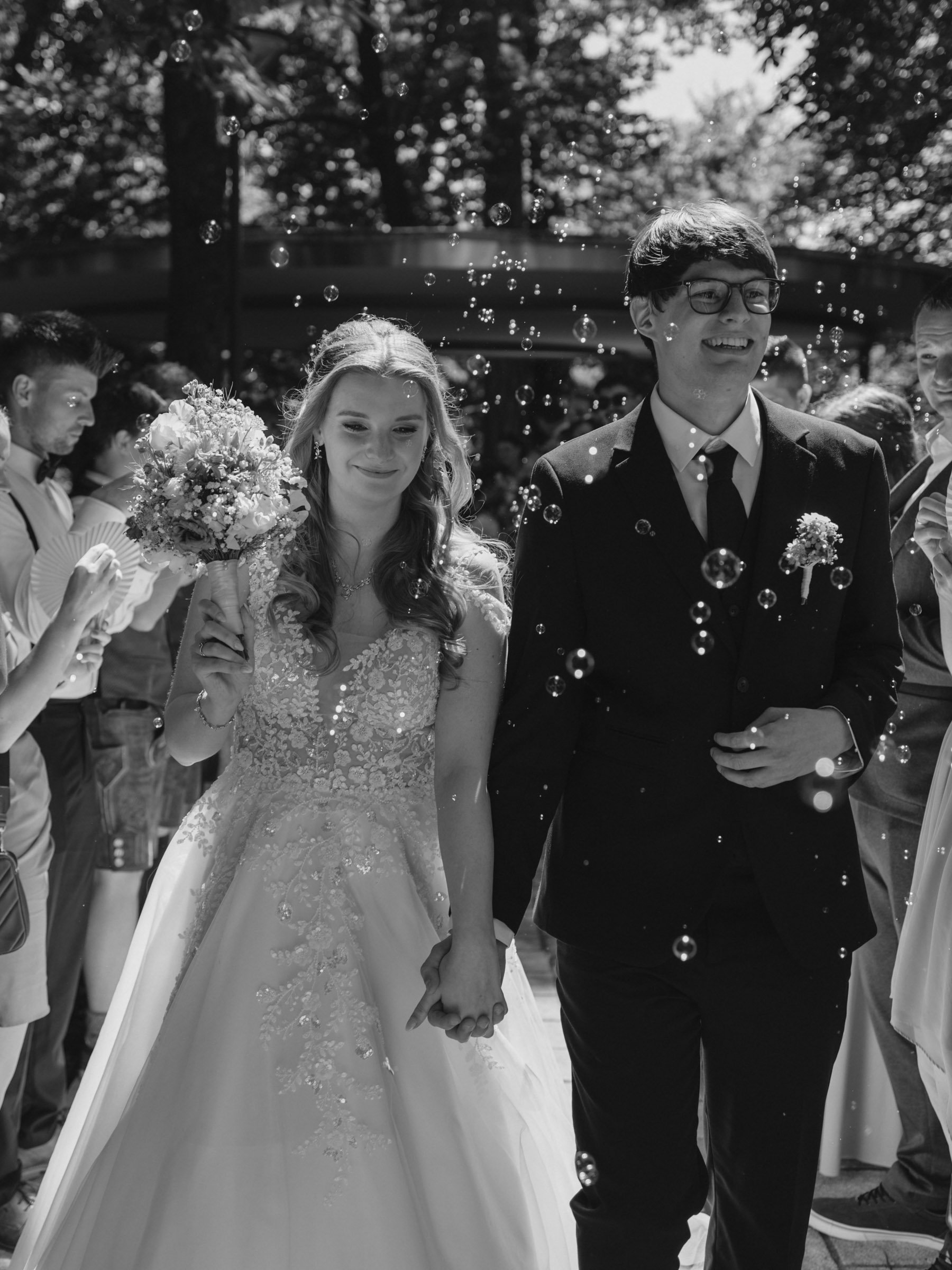 Das Bild zeigt ein Brautpaar bei einer Hochzeit, das gemeinsam die Hand hält und sich freut. Es sind noch weitere Gäste im Hintergrund, und Wasserblasen sind in der Luft sichtbar.