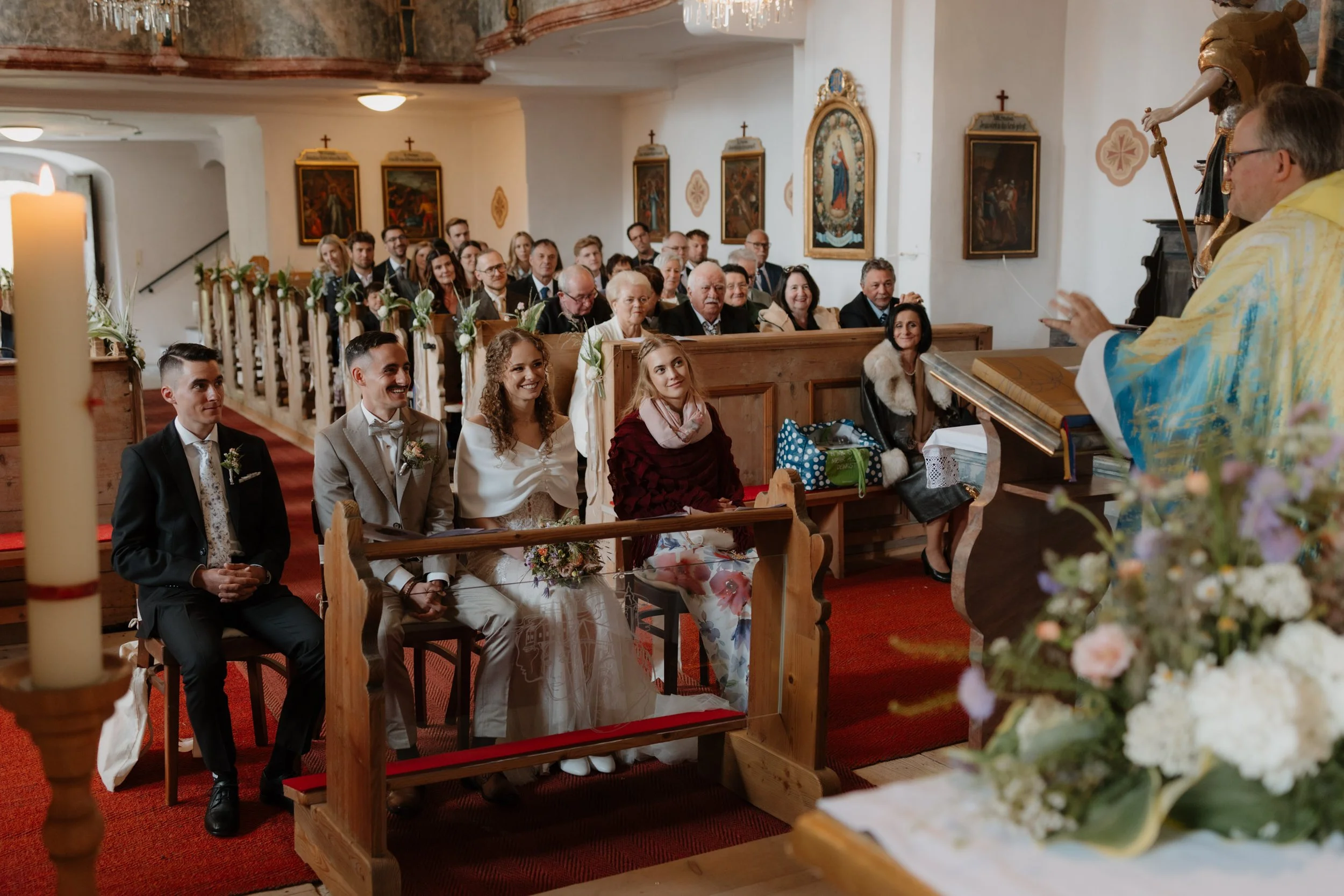 Hochzeitspaar bei der Trauung in einer Kirche, umgeben von Gästen, mit religiösen Gemälden an den Wänden.