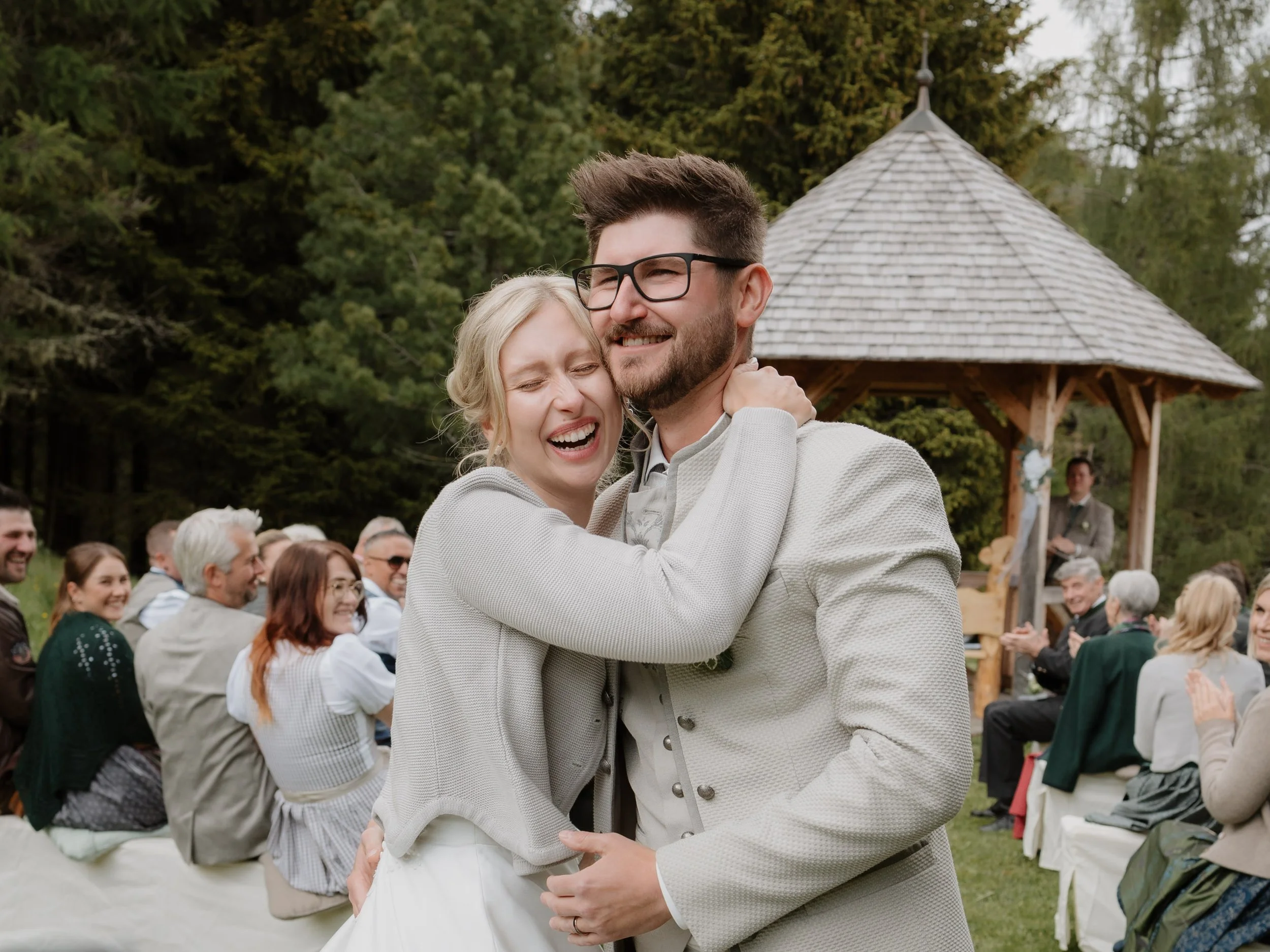 Ein lachendes Paar beim Tanzen bei einer outdoor Hochzeit, umgeben von Gästen, mit einer Holz-Architektur im Hintergrund.