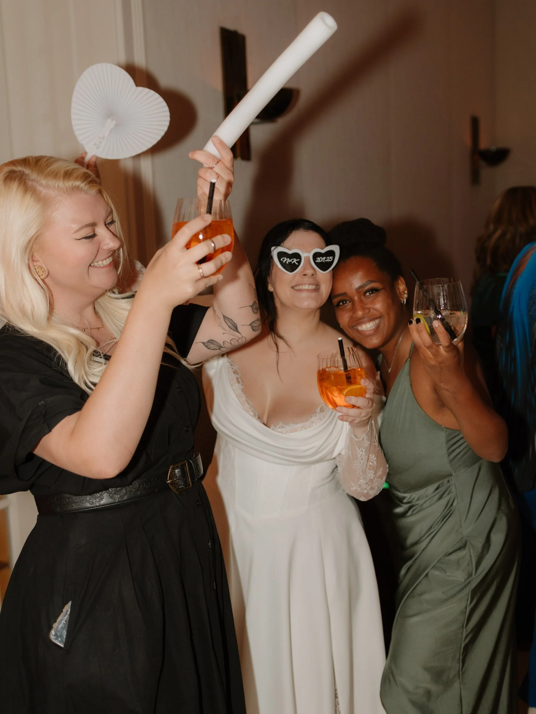 Hochzeitsparty Drei Frauen feiern zusammen bei einer Party, zwei halten Getränke, eine trägt lustige Sonnenbrille mit Schriftzügen. Sie lachen und haben Spaß.