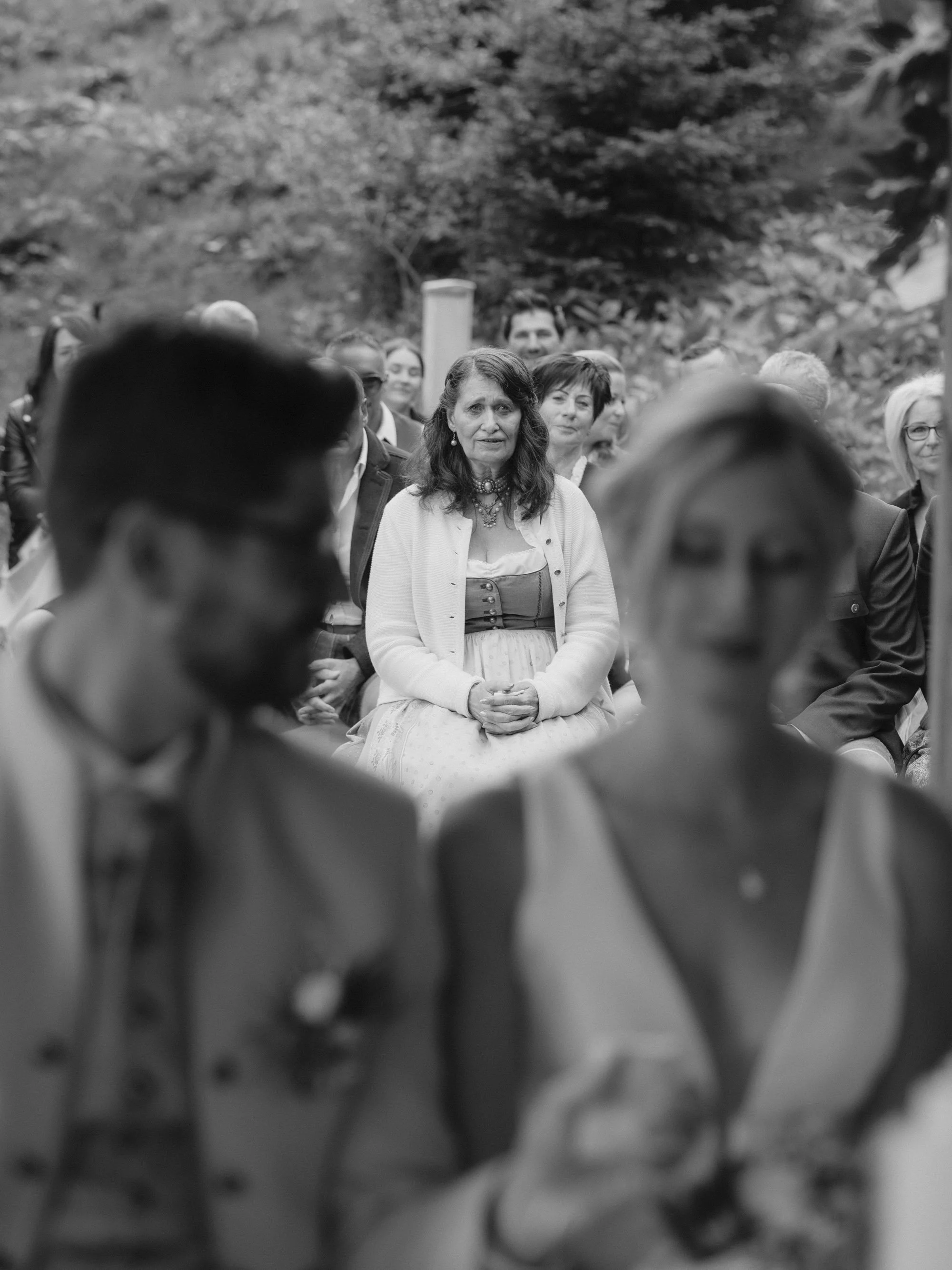 Eine Gruppe von Menschen im Freien, in der Mitte eine Frau mit dunklem Haar, die eine weiße Bluse und Schmuck trägt, sitzt und die anderen um sie herum stehen.