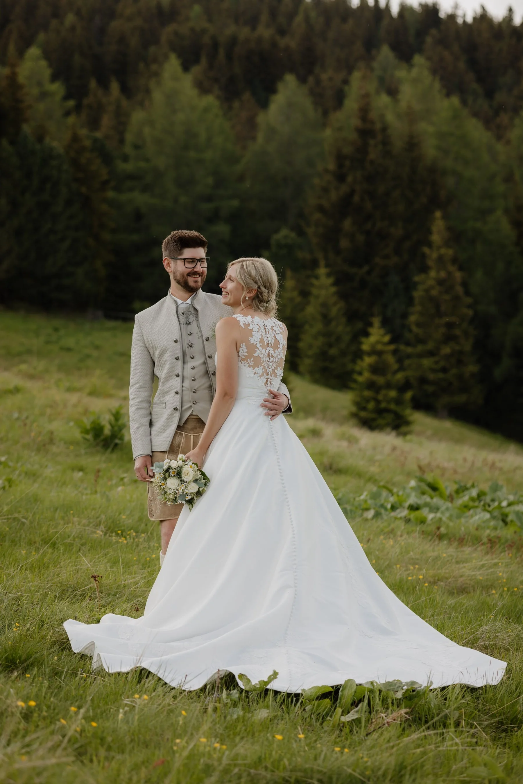 Brautpaar im Grünen, die Frau trägt ein weißes Brautkleid mit Spitze und der Mann ein graues Jackett, beide lachen sich an, im Hintergrund sind Bäume und eine Wiese.