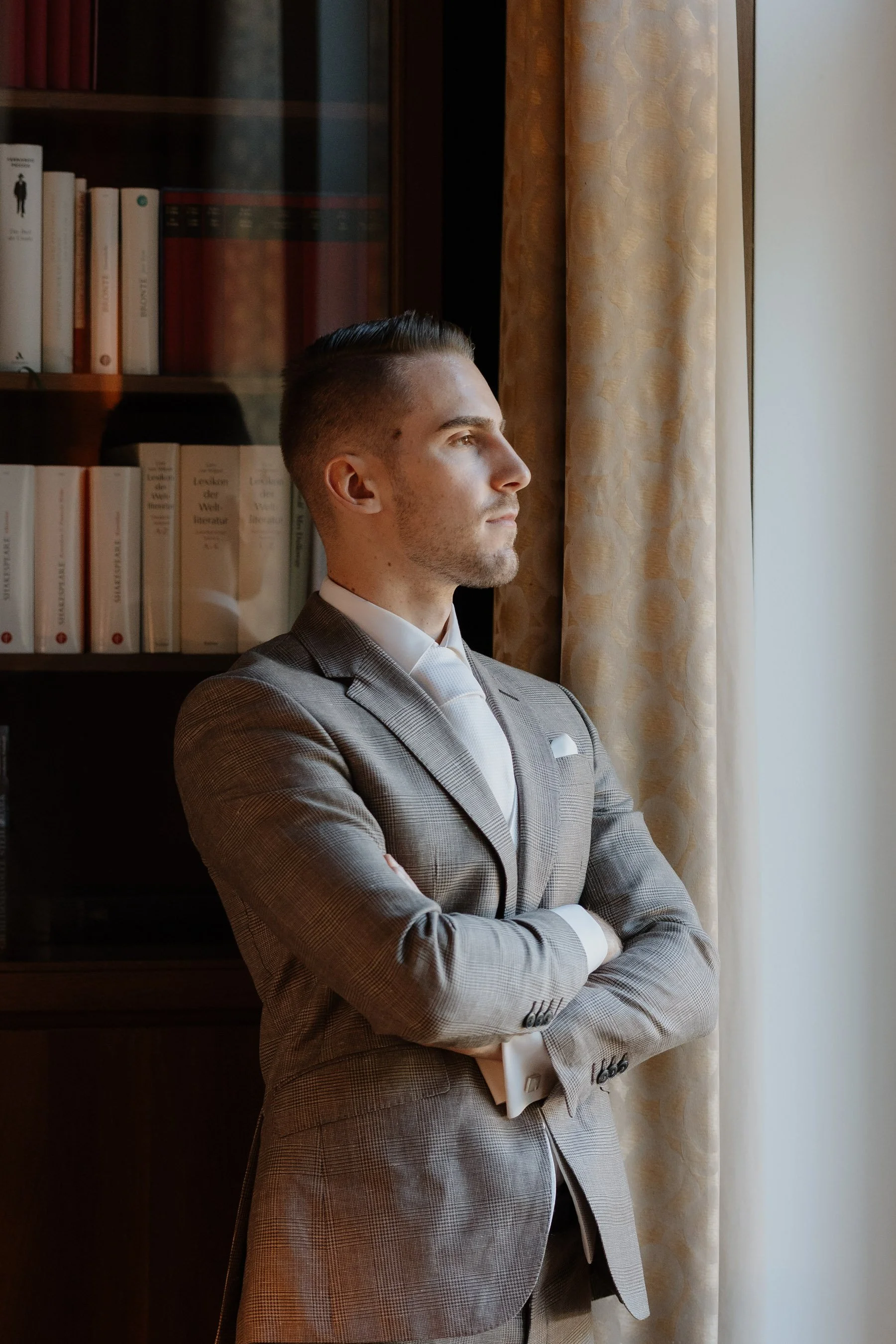 Bräutigam Portrait. Ein junger Mann in Anzug mit verschränkten Armen steht vor einer Fensterseite mit Blick nach draußen. Im Hintergrund sind Bücherregale sichtbar.