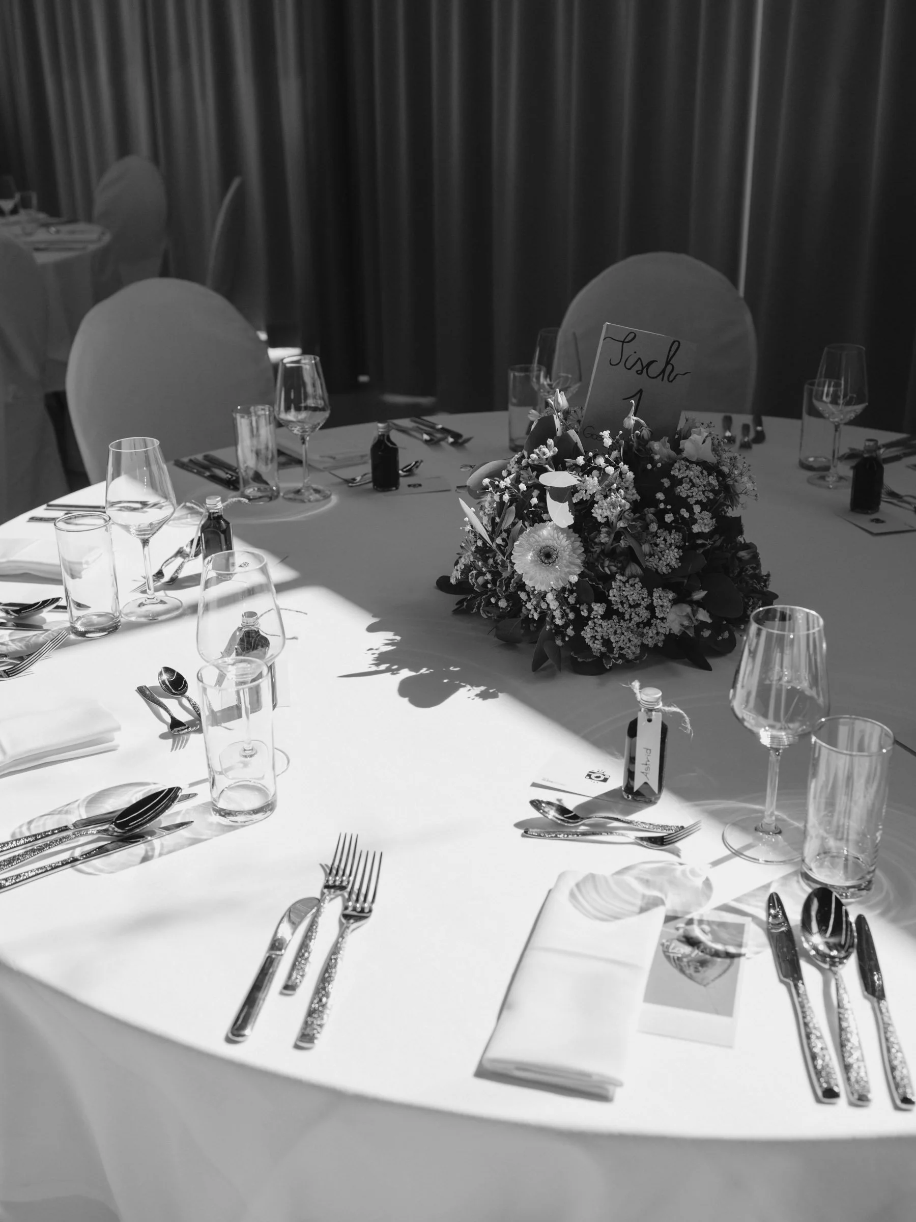 Detail Aufnahmen Saalfotos. Ein runder Esstisch ist festlich gedeckt mit Gläsern, Besteck, weißen Servietten und einer Blumenarrangement in der Mitte. Auf einem kleinen Schild steht 'Tisch 1'.