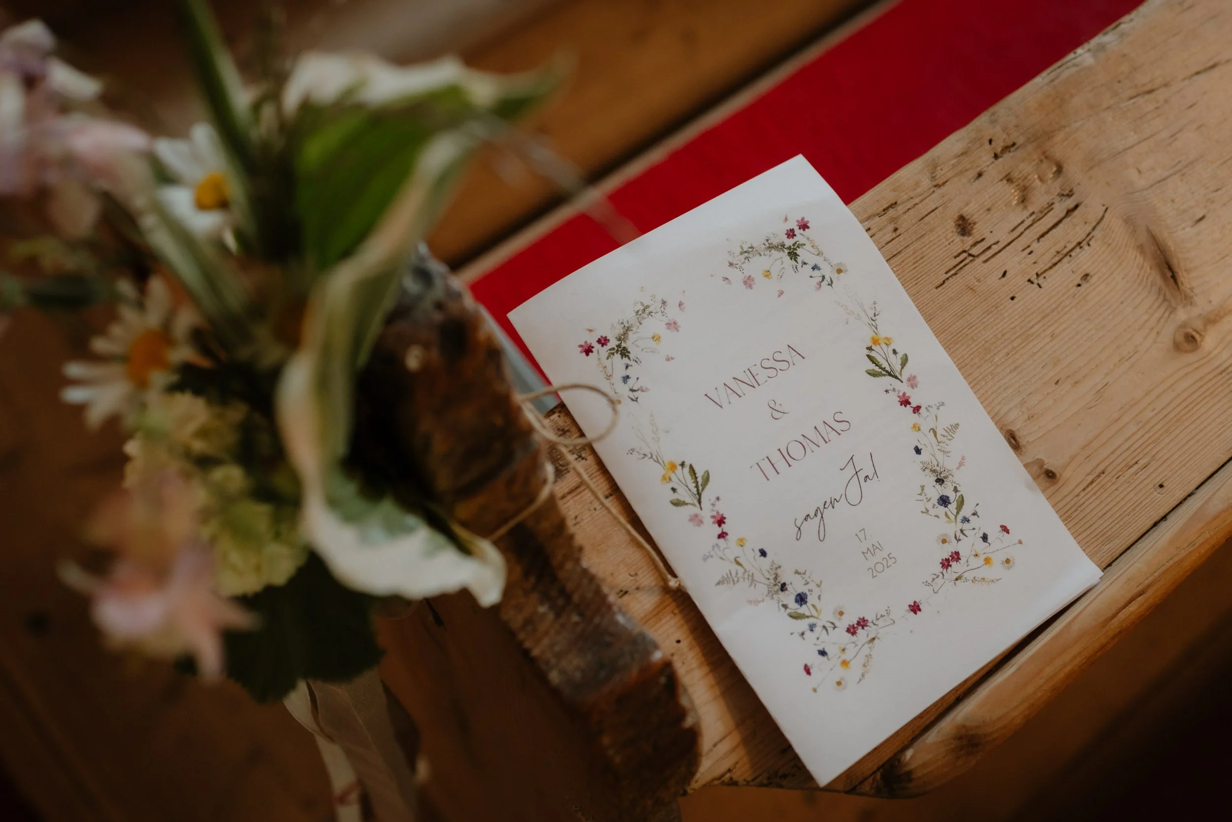 Ein Hochzeitsprogramm mit Blumenmuster auf einem Holztisch, teilweise sichtbar, neben einem Blumenstrauß mit weißen und gelben Blumen.