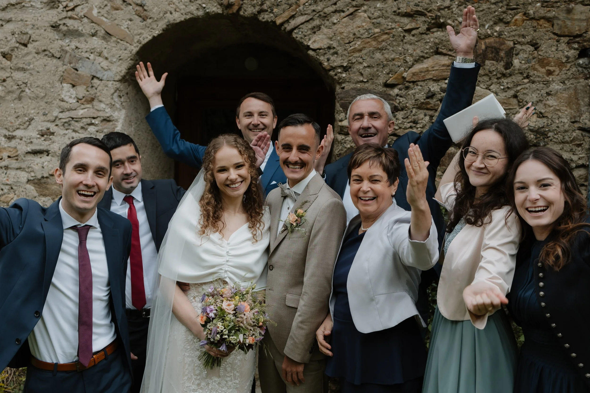 Eine Gruppe von Menschen, darunter ein Brautpaar, die bei einer Hochzeit vor einer alten Steinmauer steht, alle lächeln und feiern.
