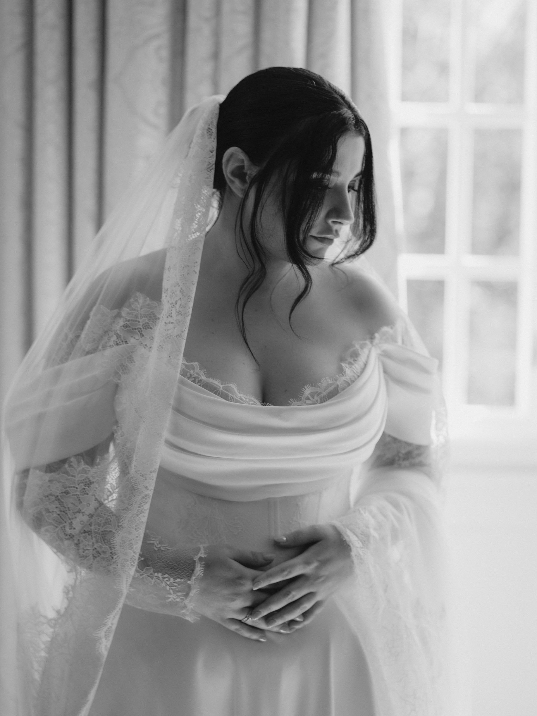 Schwarz-weiß-Fotografie einer Braut mit Brautkleid und Schleier, die ihre Hände auf ihren Bauch legt, vor einem Fenster mit Vorhängen in einem Raum.