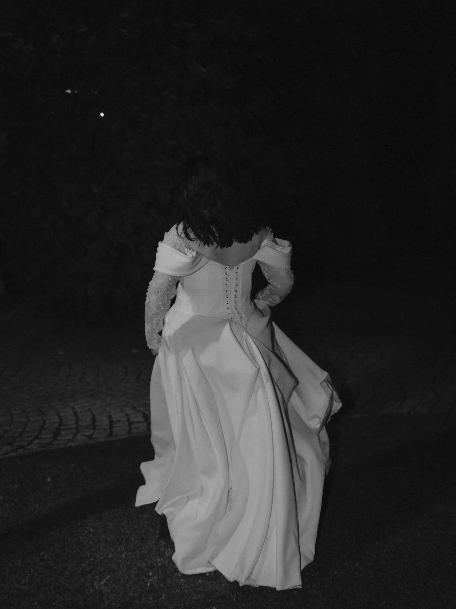 ausgefallene Hochzeitsfotos. besondere Hochzeitsfotos. Eine Person in einem langem weißen Kleid mit offener Schulter und lockigem Haar steht auf einer dunklen Straße, bei Nacht, Blick nach unten.