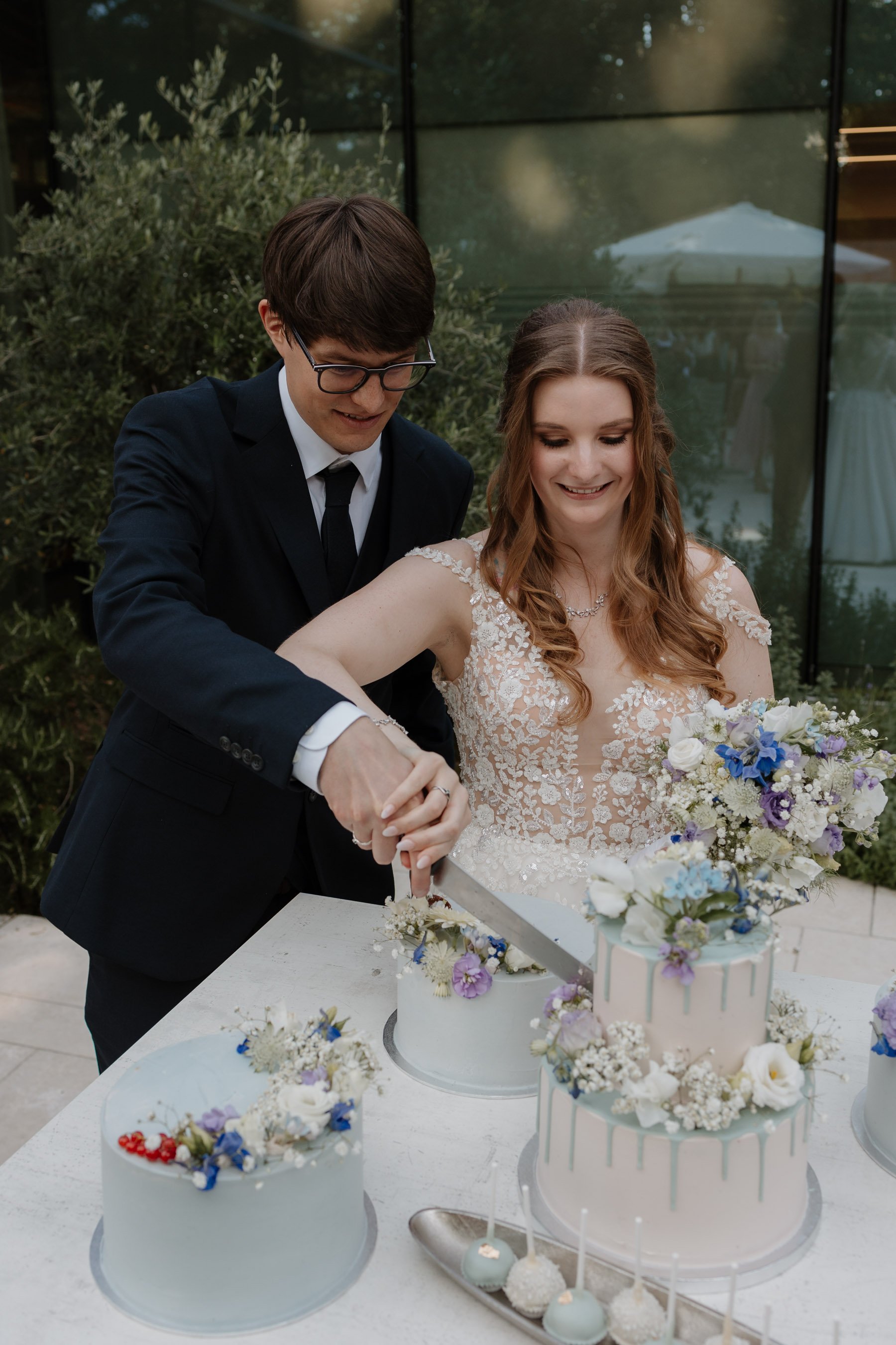 Ein Brautpaar beim Anschneiden einer Hochzeitstorte, umgeben von Blumensträußen auf einem weißen Tisch, im Hintergrund eine moderne Glasfassade und eine weiße Zelt. Die Braut trägt ein elegantes Spitzenkleid, der Bräutigam einen dunklen Anzug.
