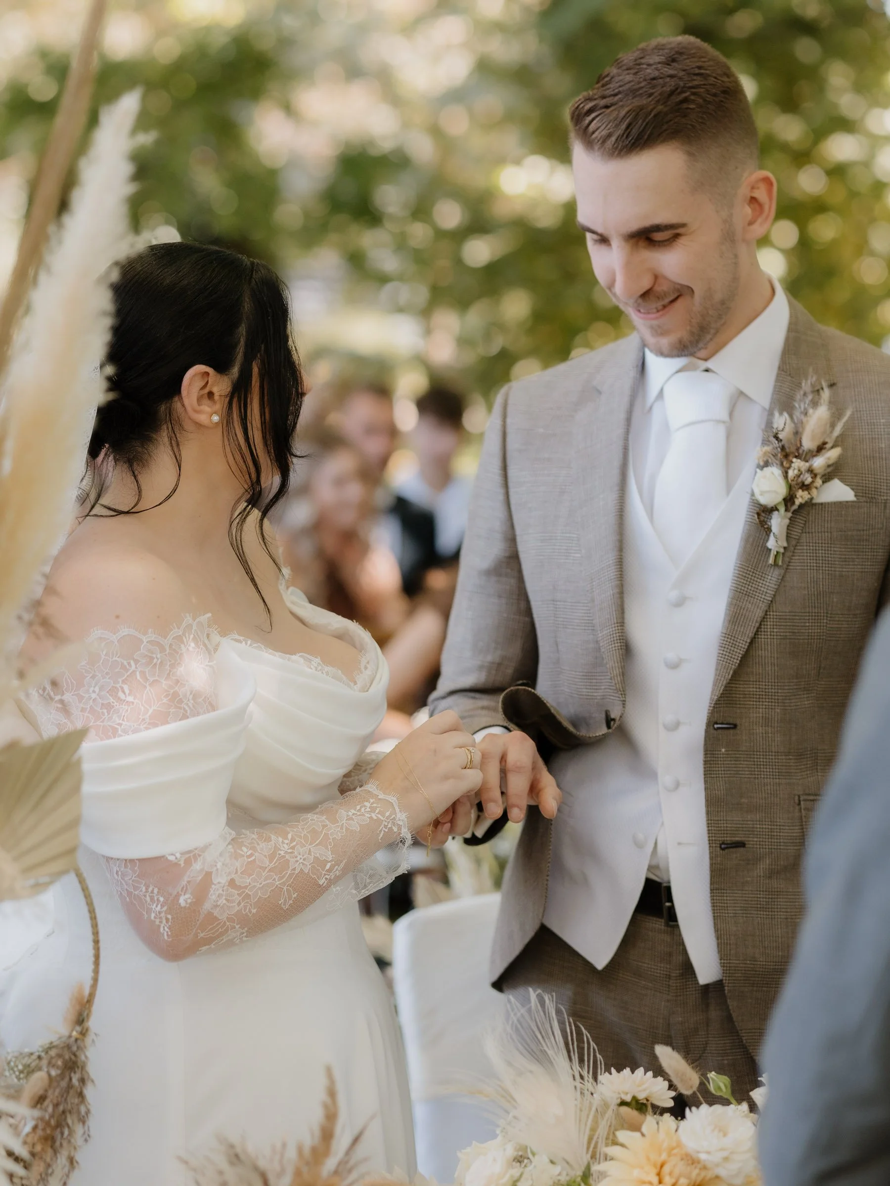 Ein Brautpaar während einer Hochzeit, das sich gegenseitig die Hand hält, im Freien bei Sonnenschein, mit Gästen im Hintergrund.