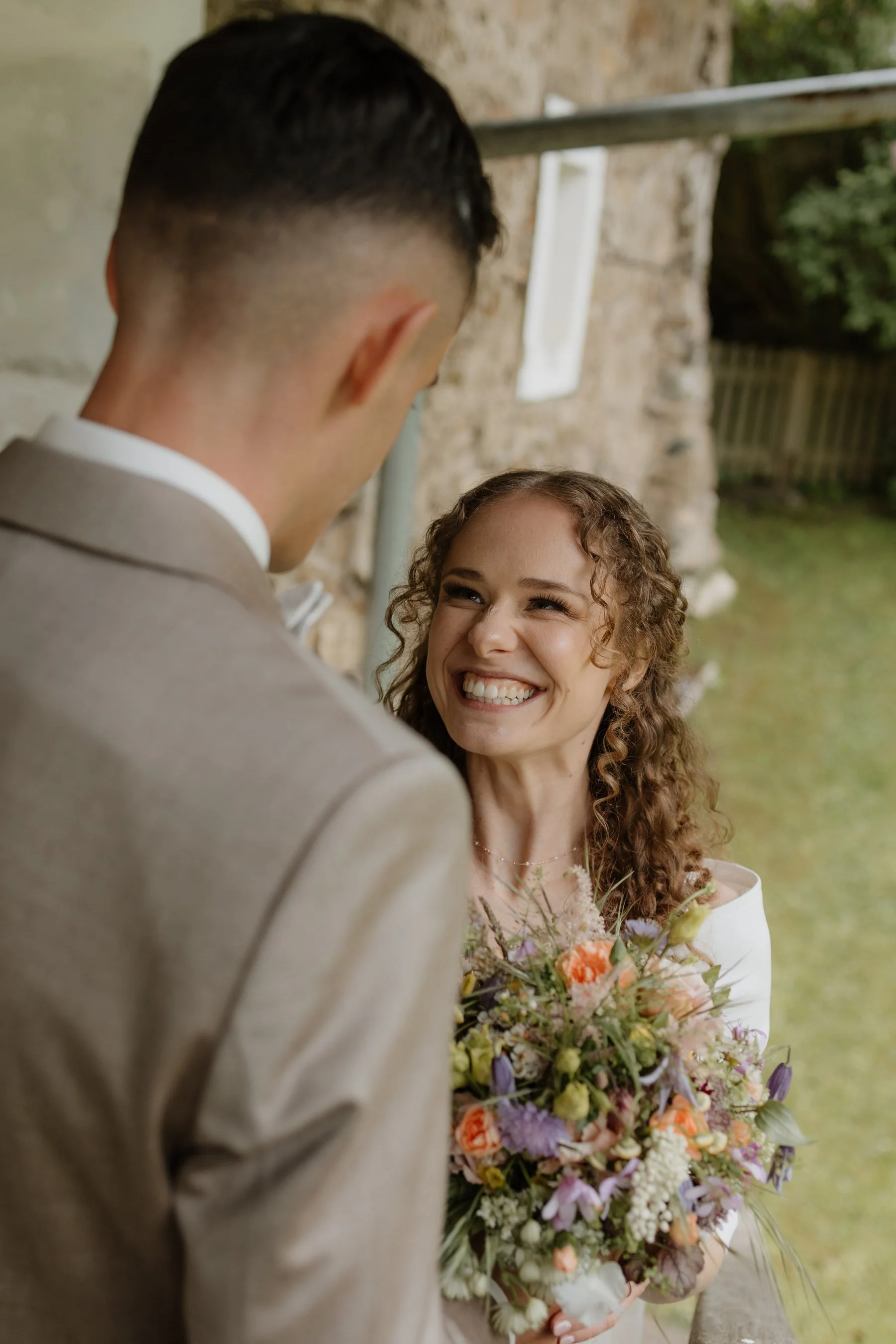 Ein braunhaariges Brautpaar bei einer Hochzeit im Freien, die Frau hält einen bunten Blumenstrauß, beide lächeln liebevoll.