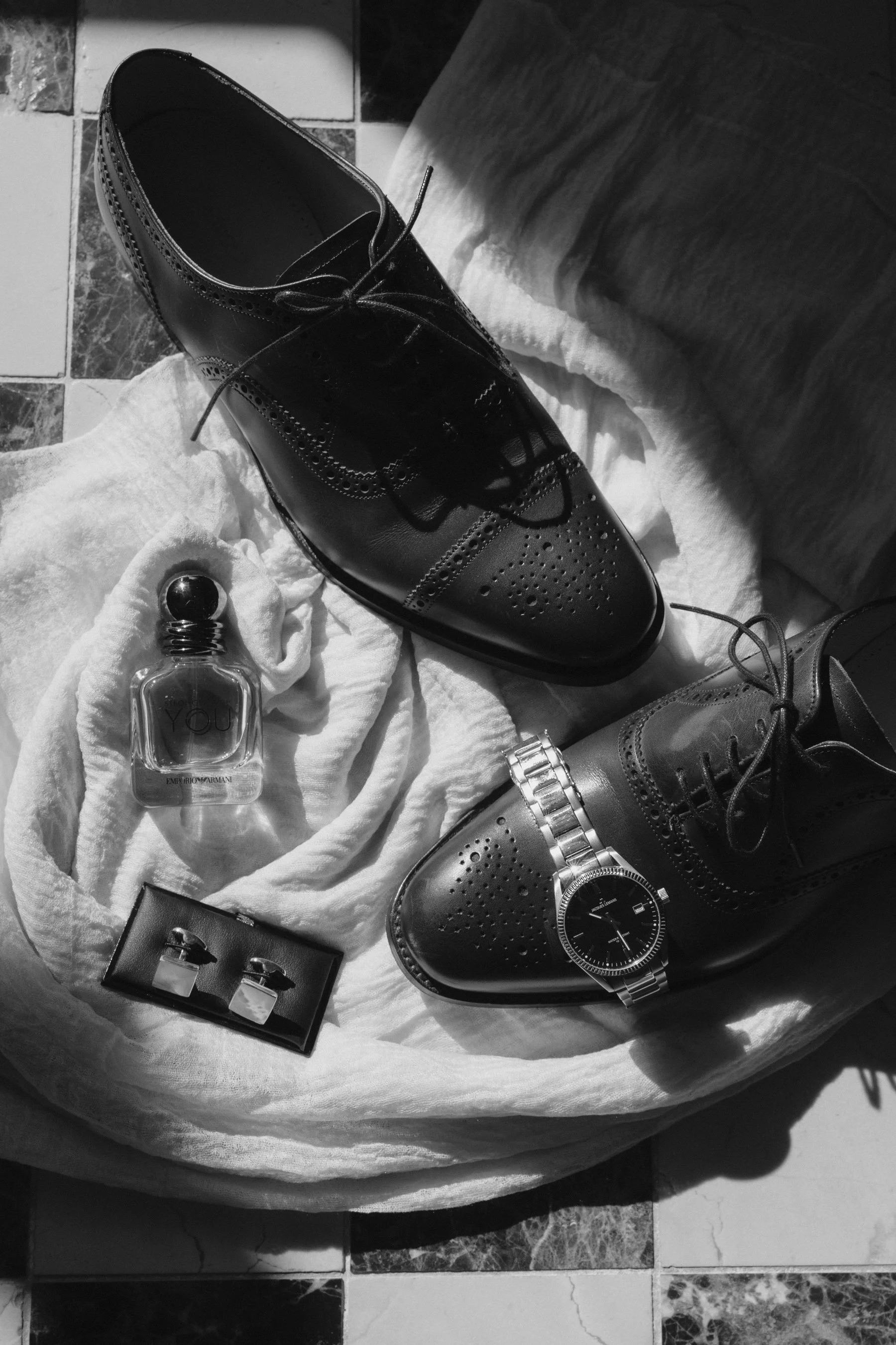 Schwarze Lederschuhe, eine Flasche Parfüm, eine silberne Armbanduhr, eine schwarze Tasche und ein schwarzes Armband auf einem weißen Stofftuch, mit Marmor- und Fliesenhintergrund.