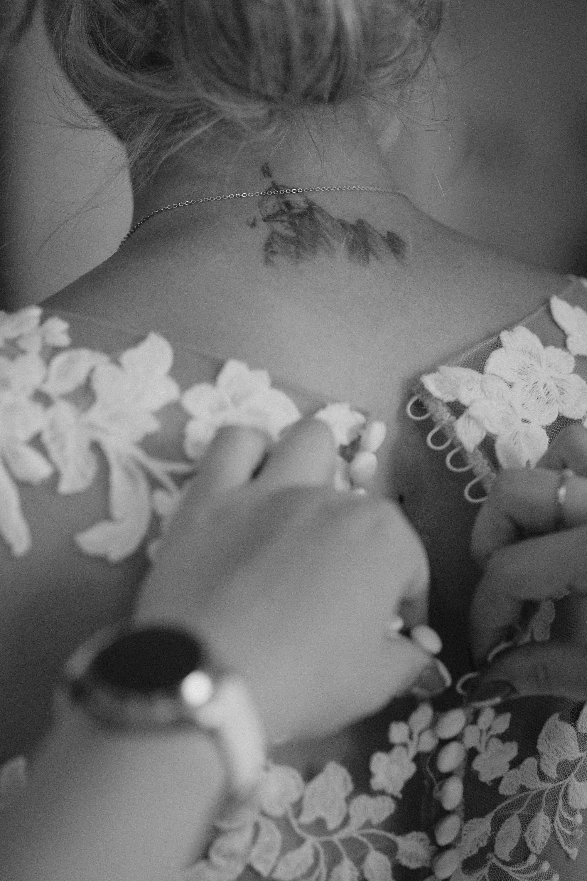 Nahaufnahme eines nackten Rückens mit Tattoo und weißen Blumen, dunkle Haare, silberne Halskette, schwarze und weiße Kleidung.