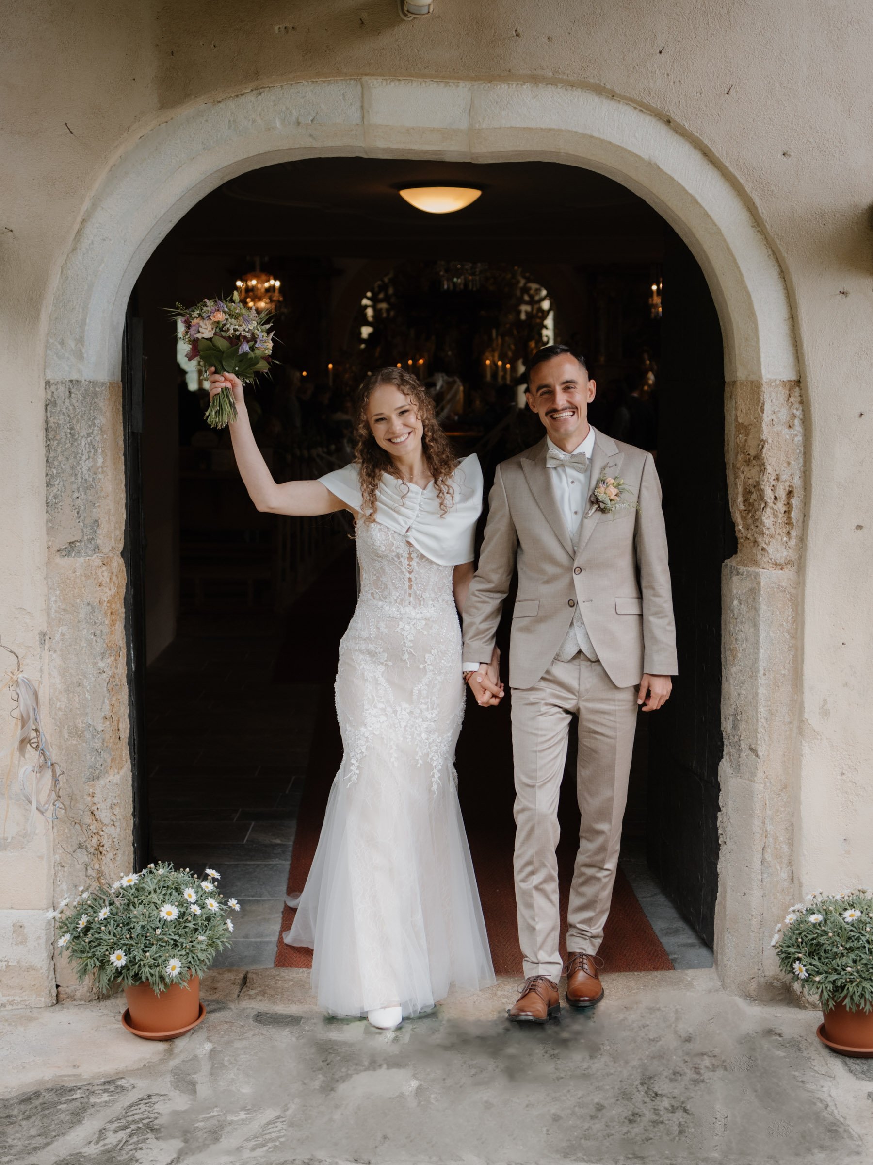 Ein glückliches Brautpaar beim Verlassen einer Kirche, die Braut trägt ein weißes Hochzeitskleid und hält einen Blumenstrauß hoch, der Bräutigam trägt einen beige Anzug mit Fliege. Sie halten sich an den Händen und lächeln.
