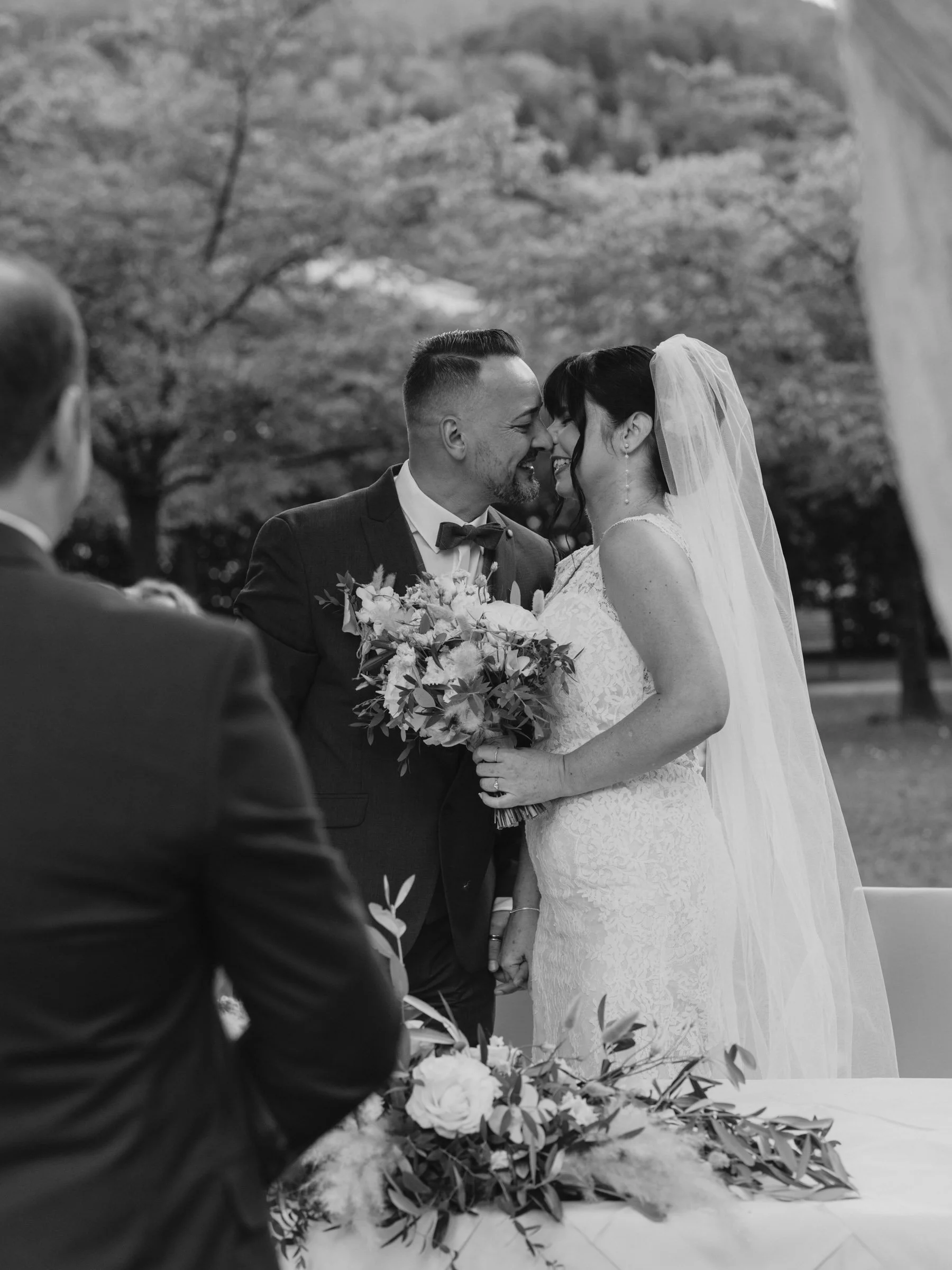 Ein Brautpaar bei ihrer Hochzeit, Nahaufnahme auf das gemeinsame Lächeln, im Hintergrund Bäume, schwarz-weiß Fotografie, romantische Atmosphäre.