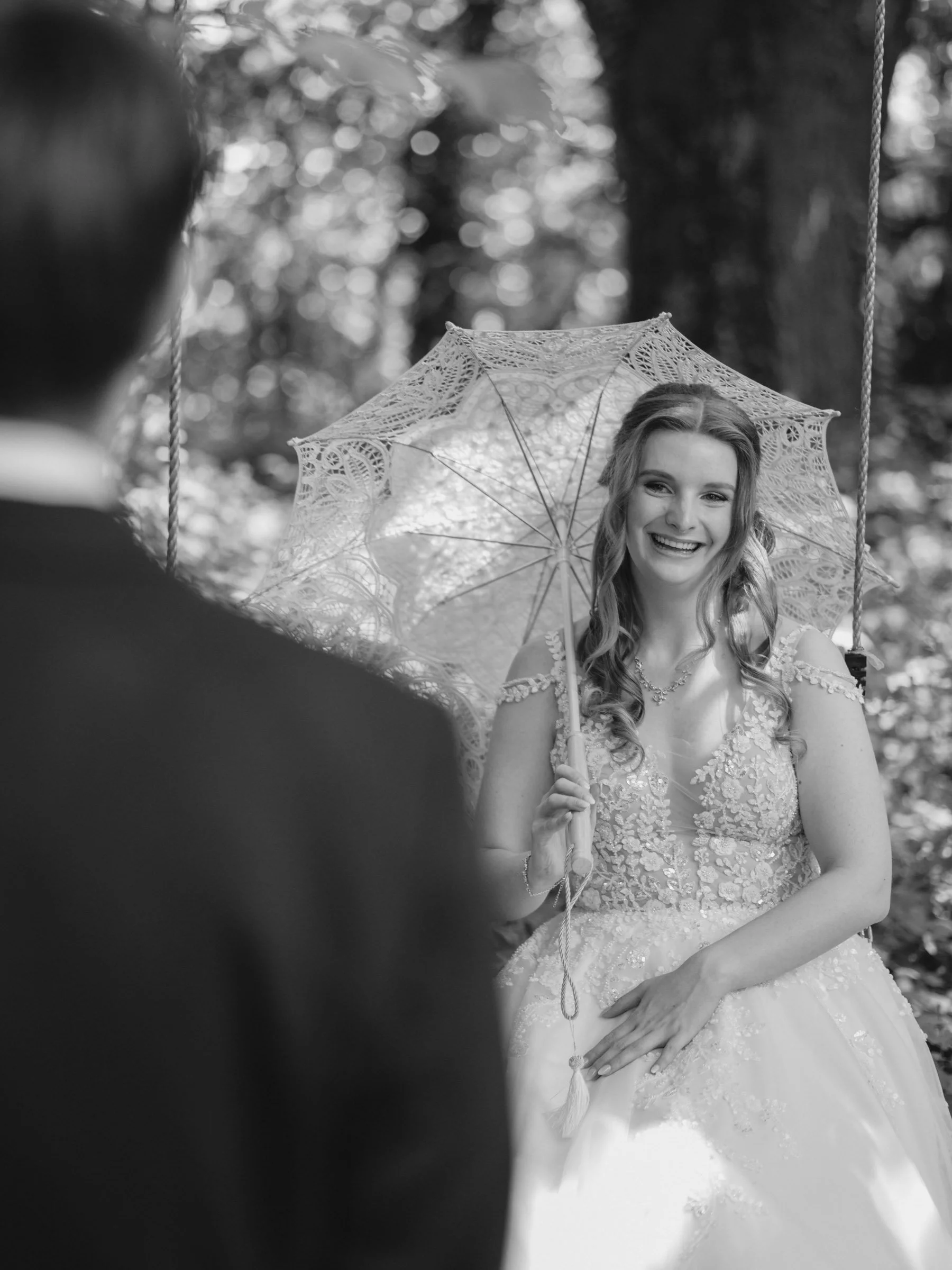 Eine glückliche Frau in einem weißen Brautkleid, die eine Spitze-Regenschirm hält, wird bei einer Hochzeit im Freien fotografiert. Ein Mann in dunkler Kleidung ist im Vordergrund unscharf.