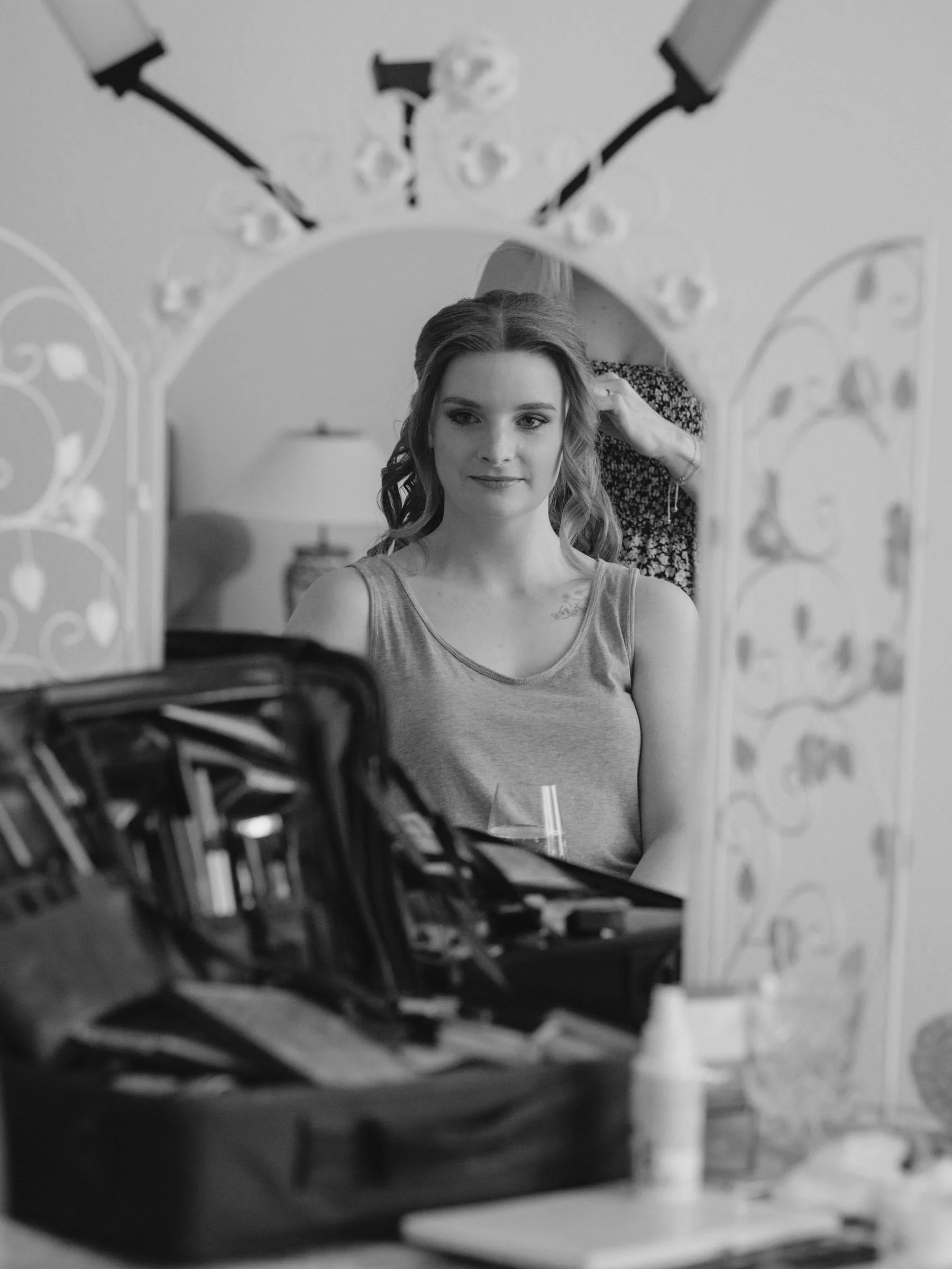 Getting Ready der Braut. Ein junges Mädchen mit langem Haar sitzt vor einem Spiegel, während eine Person im Hintergrund ihre Haare frisiert. Das Bild ist schwarz-weiß.