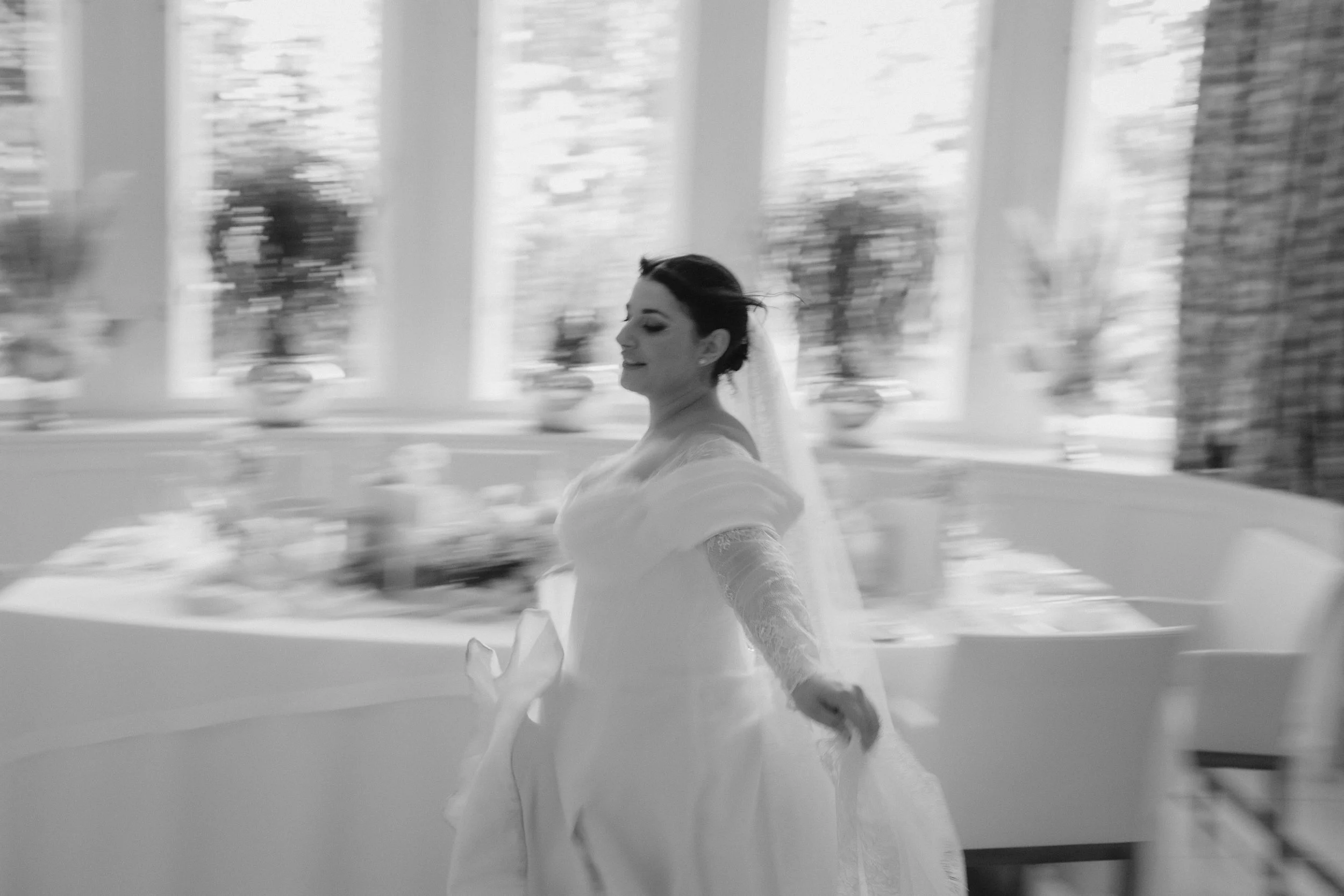 besondere Hochzeitsfotos, ausgefallen, einzigartige Hochzeitsfotos. Braut in einem weißen Brautkleid mit geschlossenen Augen, im Hintergrund große Fenstertüren mit Blick nach draußen, fotografiert im Schwarz-Weiß-Stil.