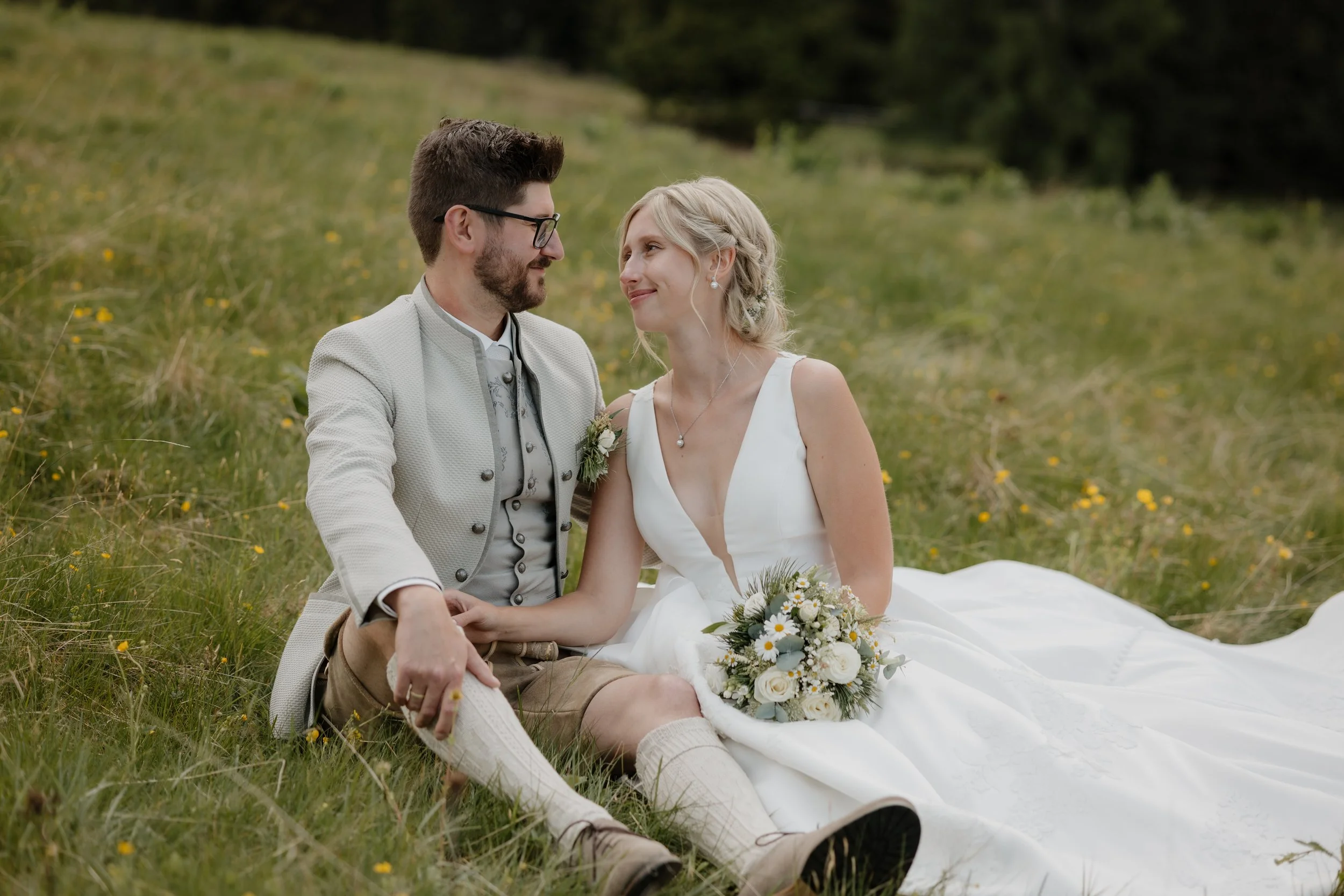 Ein Brautpaar sitzt verliebt in einem grünen Feld mit kleinen gelben Blumen, trägt Hochzeitskleidung und schaut sich an.