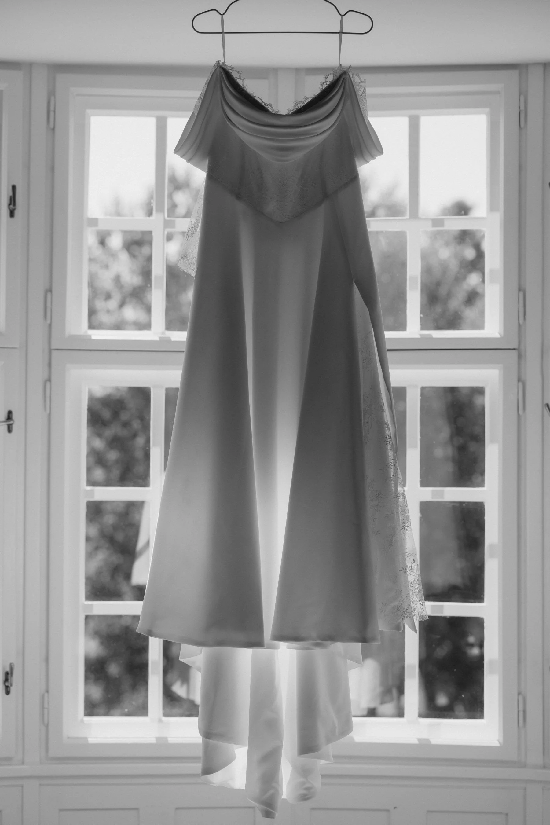 Detailaufnahmen Hochzeit. Ein weißes Brautkleid und ein Anzug hängen an einem Holzkleiderbügel vor einem Fenster.