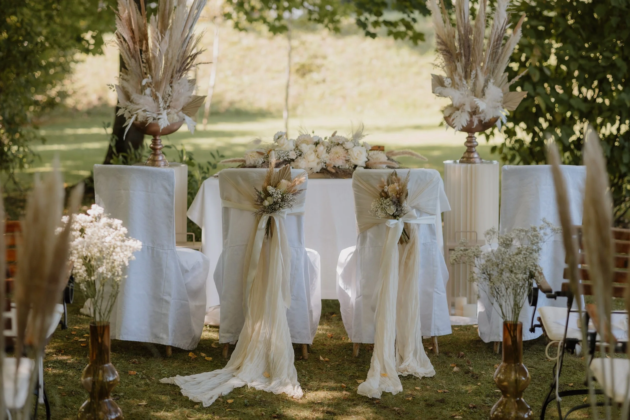 Eine elegante Hochzeitstafel im Freien mit weißen Tischtüchern, Blumenarrangements in Weiß- und Beigetönen, großformatigen Dekorationen mit Federn und Trockenblumen, und schlichten weißen Stühlen