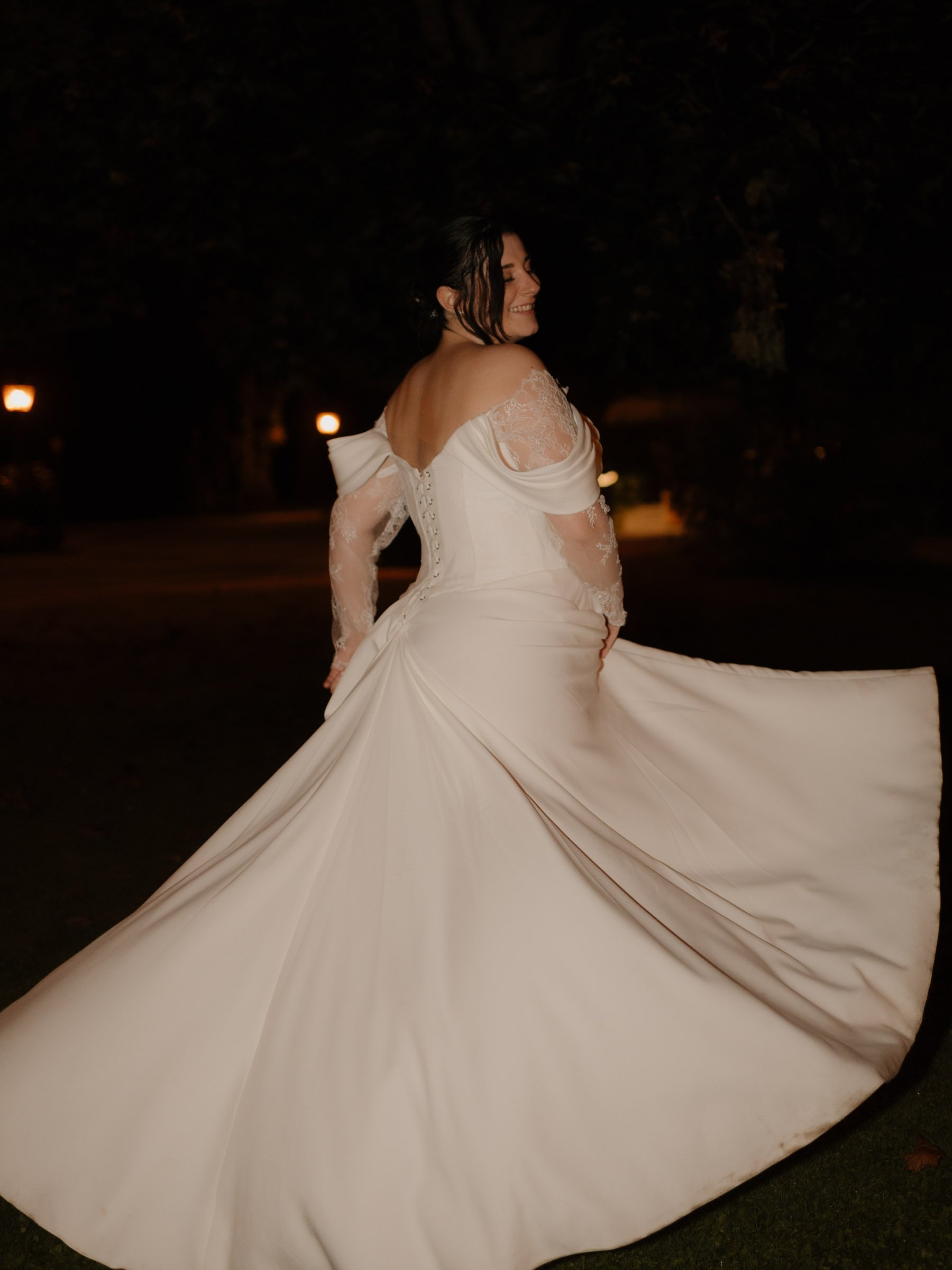 ausgefallene Hochzeitsfotos. besondere Hochzeitsfotos. Eine lächelnde Frau trägt ein weißes Brautkleid mit langen, lace-Bässen, im Dunkeln bei Nacht, umgeben von Bäumen und Laternenlichtern.