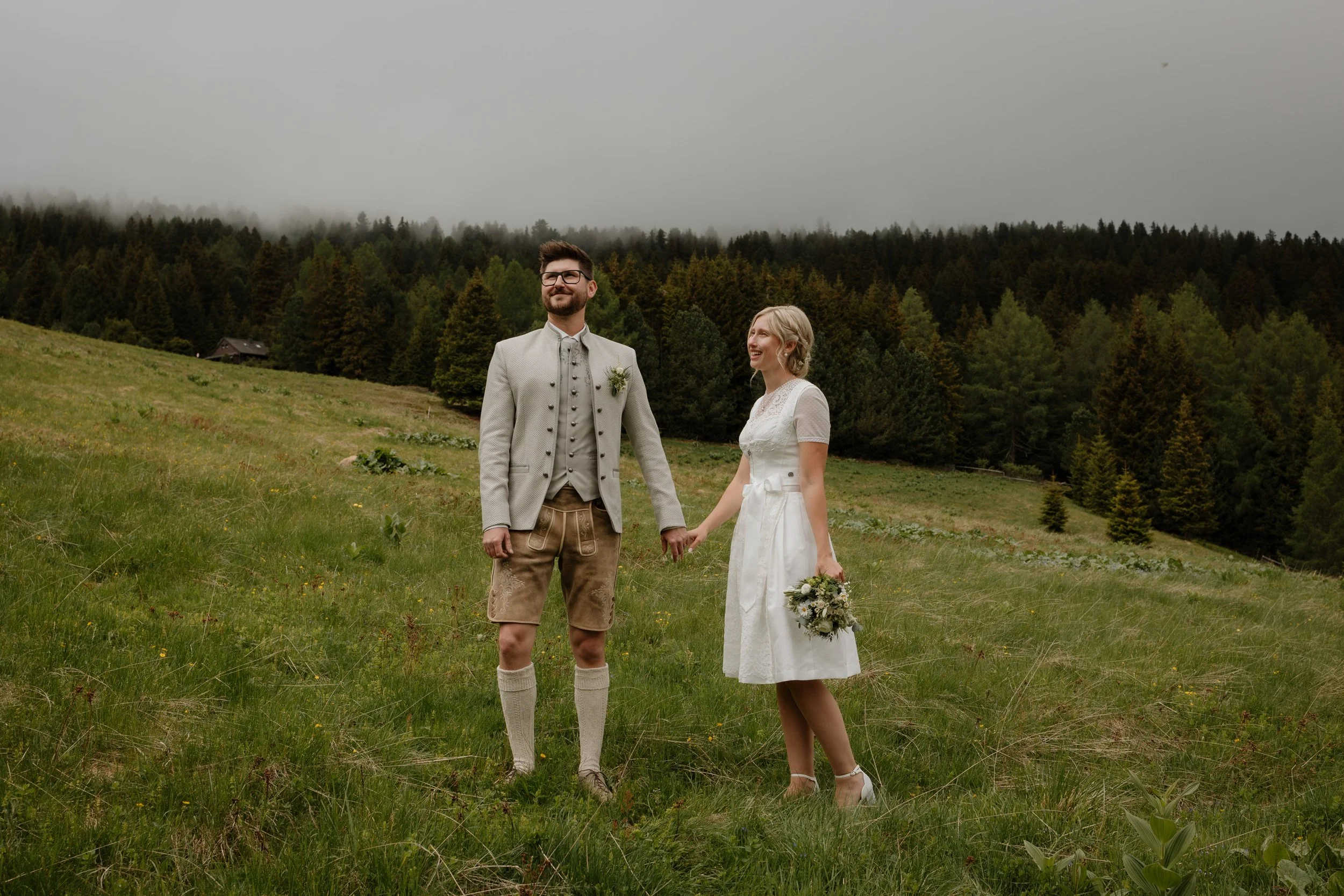 Ein Paar bei einer Hochzeit im Freien, sie halten Händchen, die Frau trägt ein weißes Hochzeitskleid, der Mann traditionelle Trachtenkleidung, im Hintergrund ein bewölkter Himmel und eine ruhige, bewaldete Landschaft.