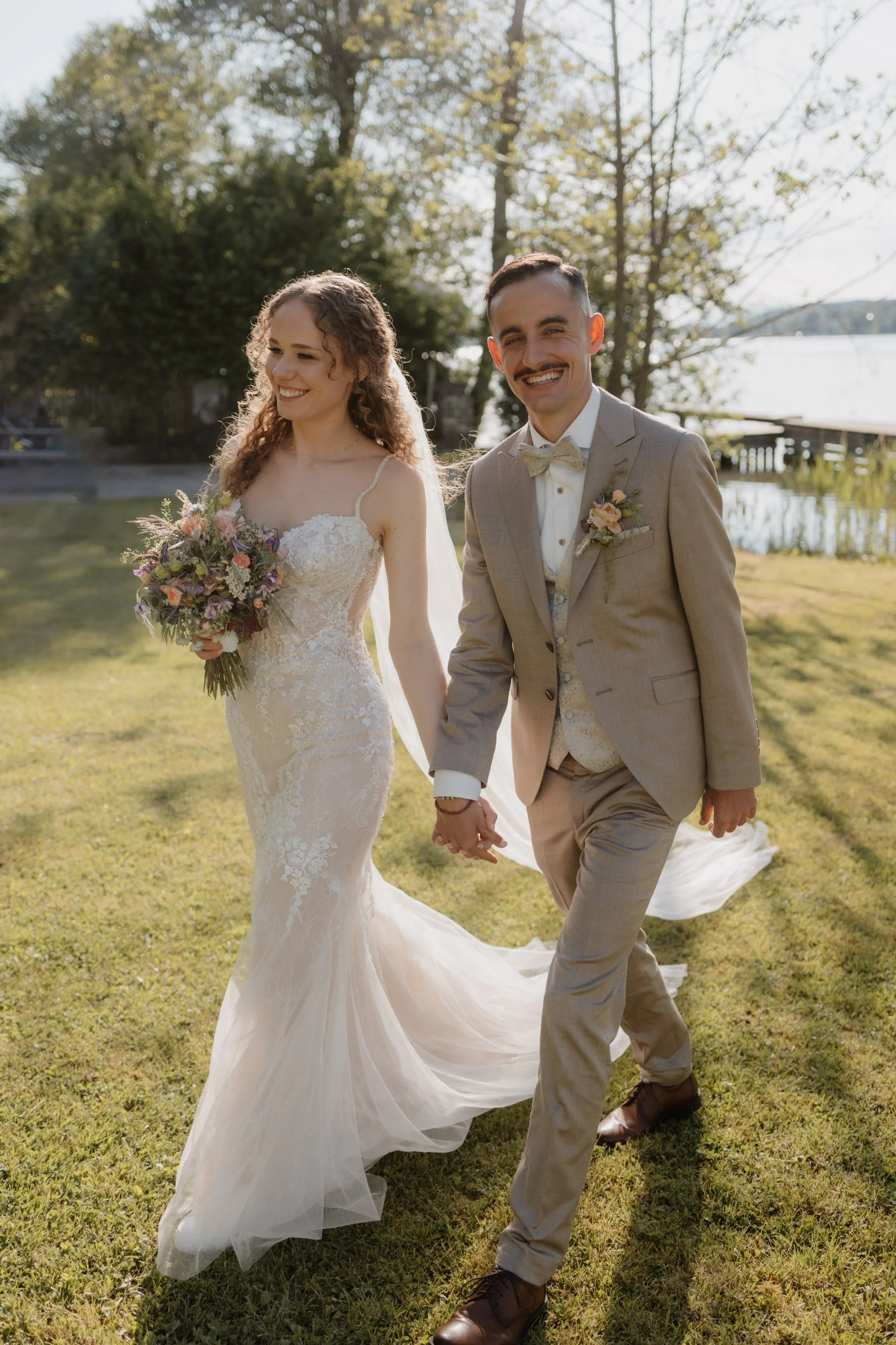 Ein frisch vermähltes Paar läuft Hand in Hand am See entlang, die Frau trägt ein langes, weißes Brautkleid und hält einen Blumenstrauß, der Mann trägt einen hellgrauen Anzug mit Fliege, im Hintergrund sind Bäume und Wasser sichtbar, die Sonne scheint