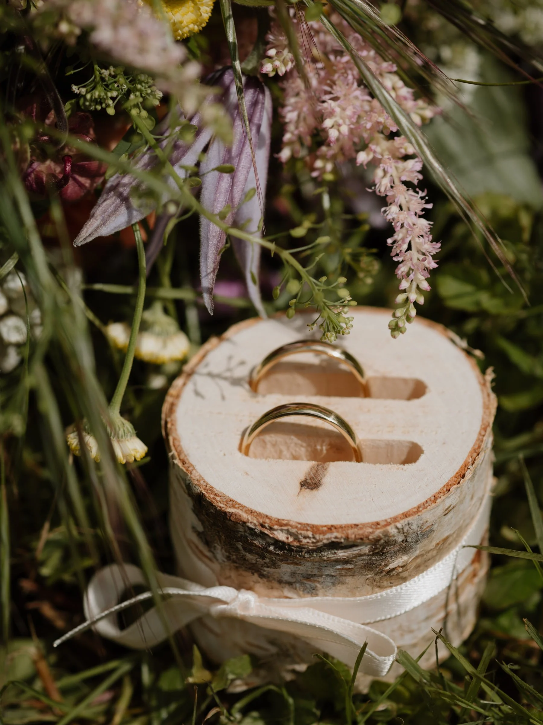  Eheringe, umgeben von Blumen, Gräsern und einem weißen Band, das auf einer grünen Wiese liegt.