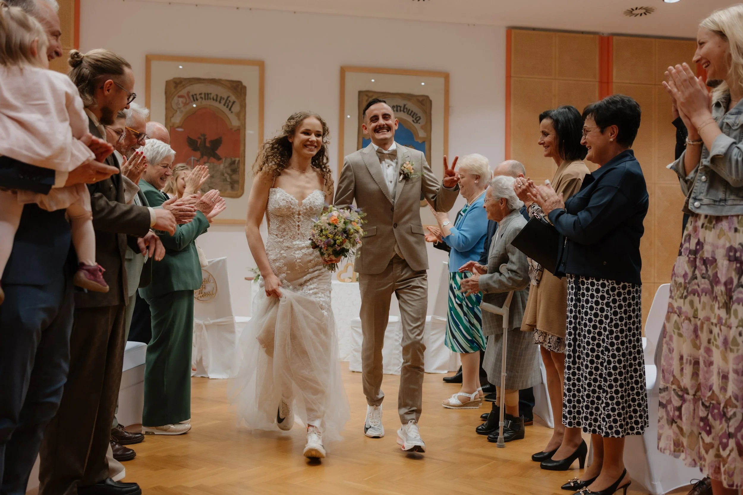 Ein Brautpaar läuft lachend durch eine versammelte Gruppe von Gästen bei einer Hochzeit. Beide sind glücklich und die Gäste applaudieren und feiern sie.