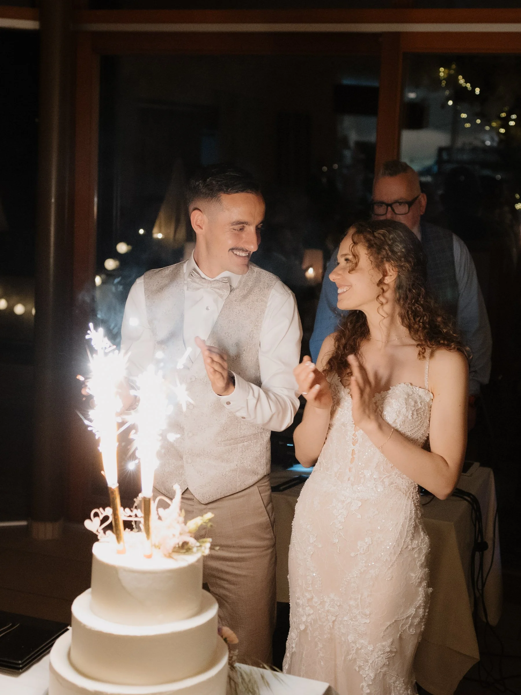 Ein Brautpaar bei ihrer Hochzeit mit einer Kerze im Vordergrund und einem Hochzeitstorte, beide lächeln sich an.