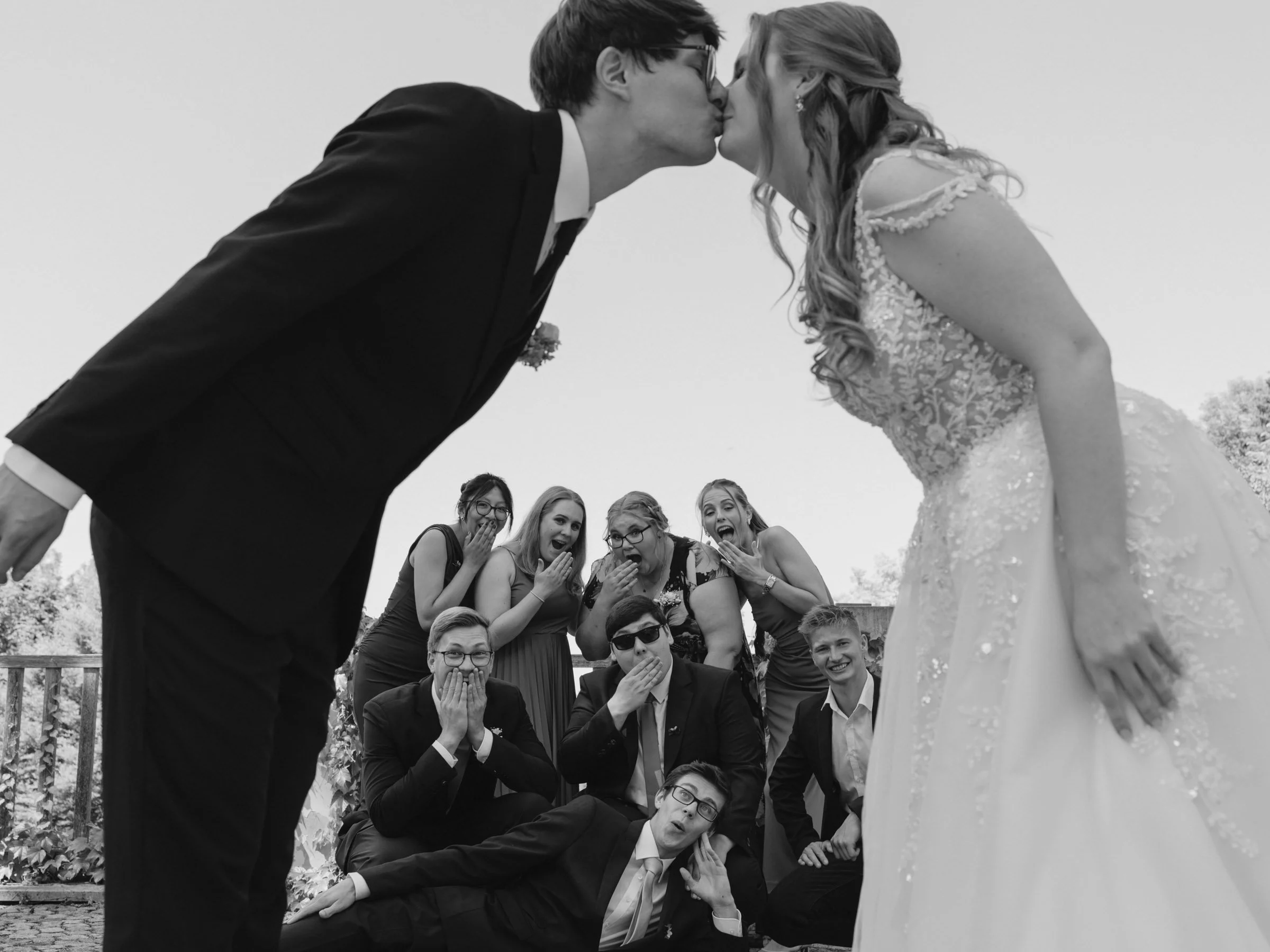 Ein Brautpaar küsst sich, während eine Gruppe von Hochzeitsgästen im Hintergrund überrascht und erfreut schaut, bei einer Hochzeit im Freien.