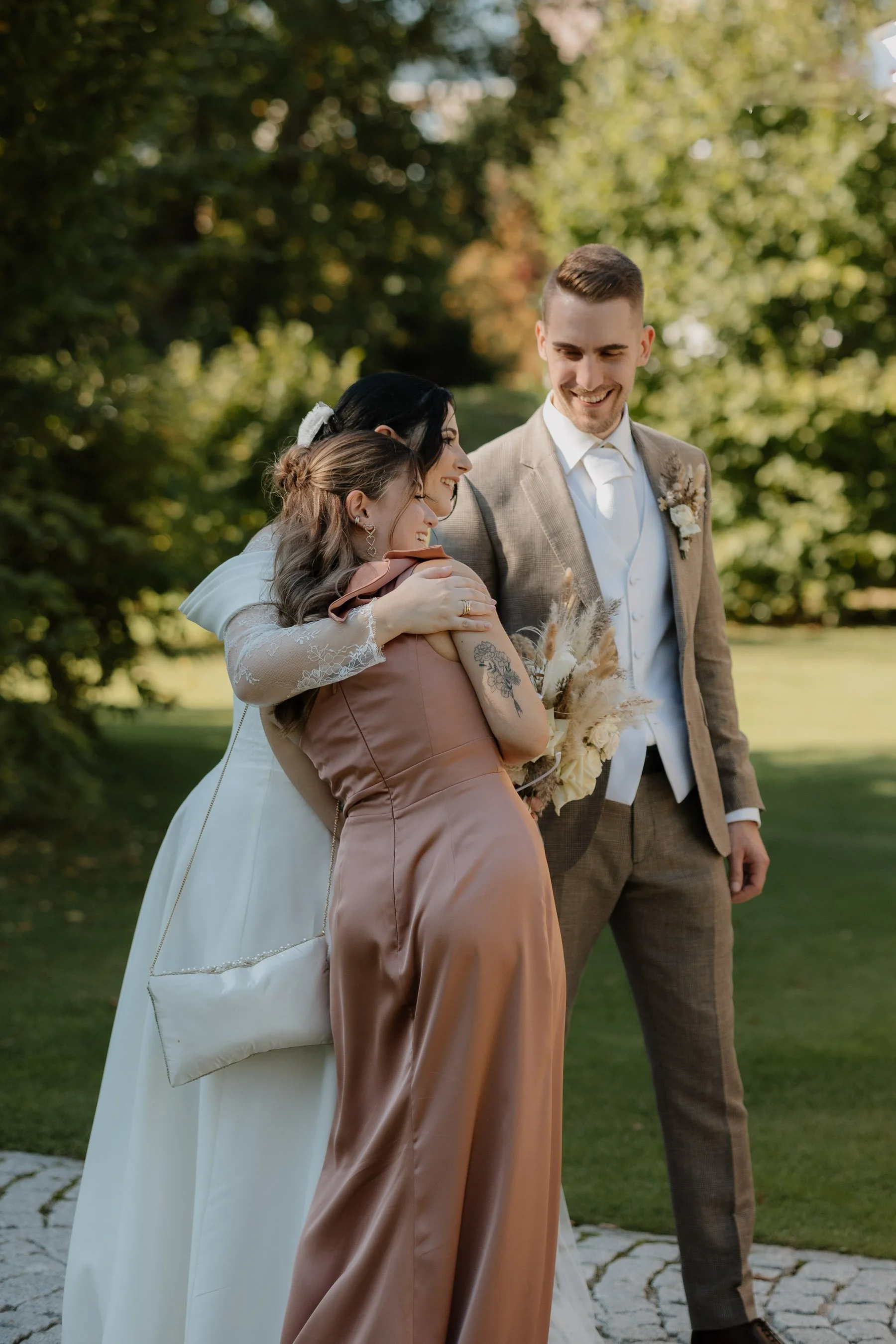 Drei Menschen beim Händeschütteln im Freien, vermutlich bei einer Hochzeit, umgeben von Bäumen, freundliches Lächeln, elegante Kleidung.