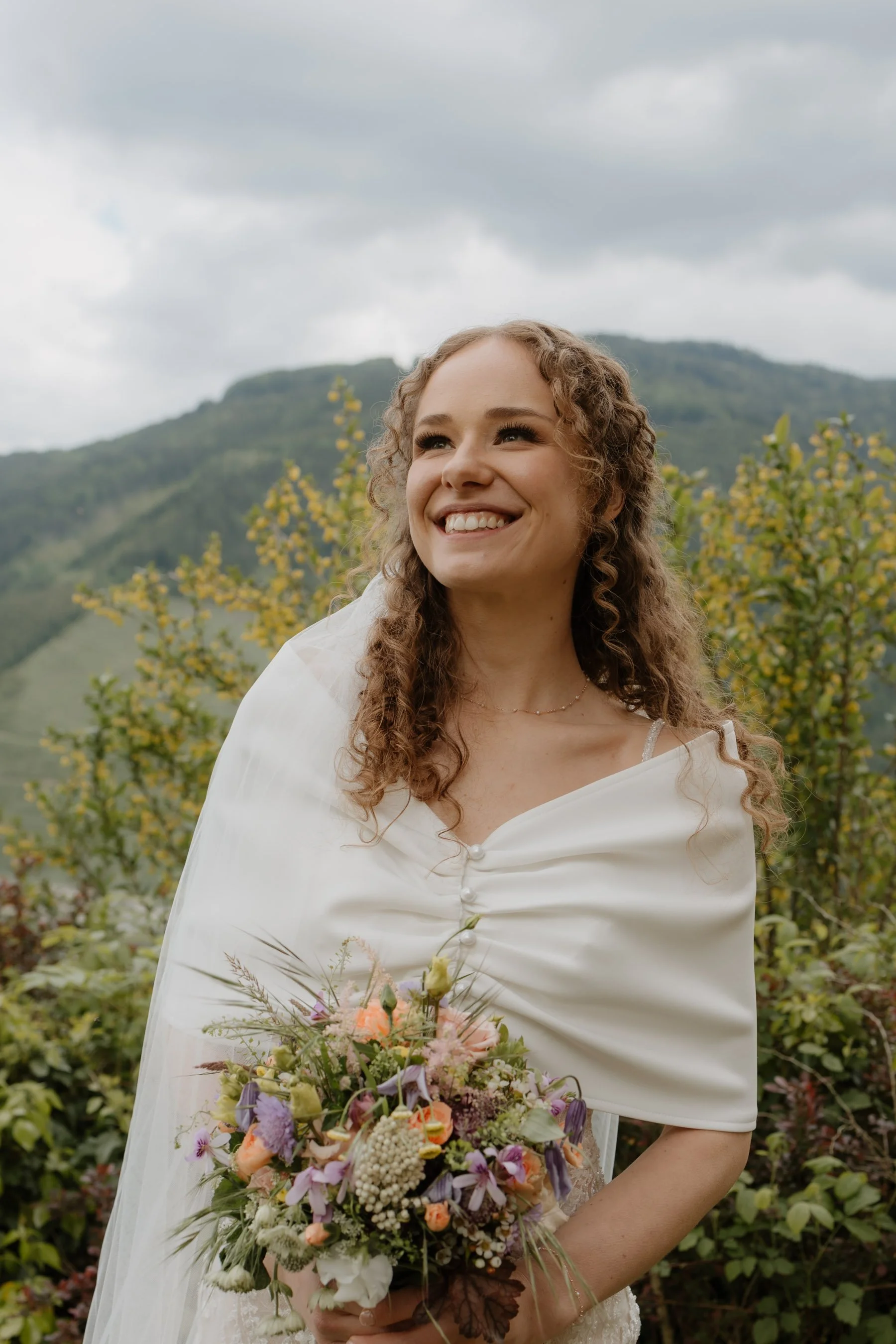 Eine lachende junge Frau im Brautkleid mit Blumenstrauß bei bewölktem Himmel, im Freien vor grünen Büschen und Bergen im Hintergrund.