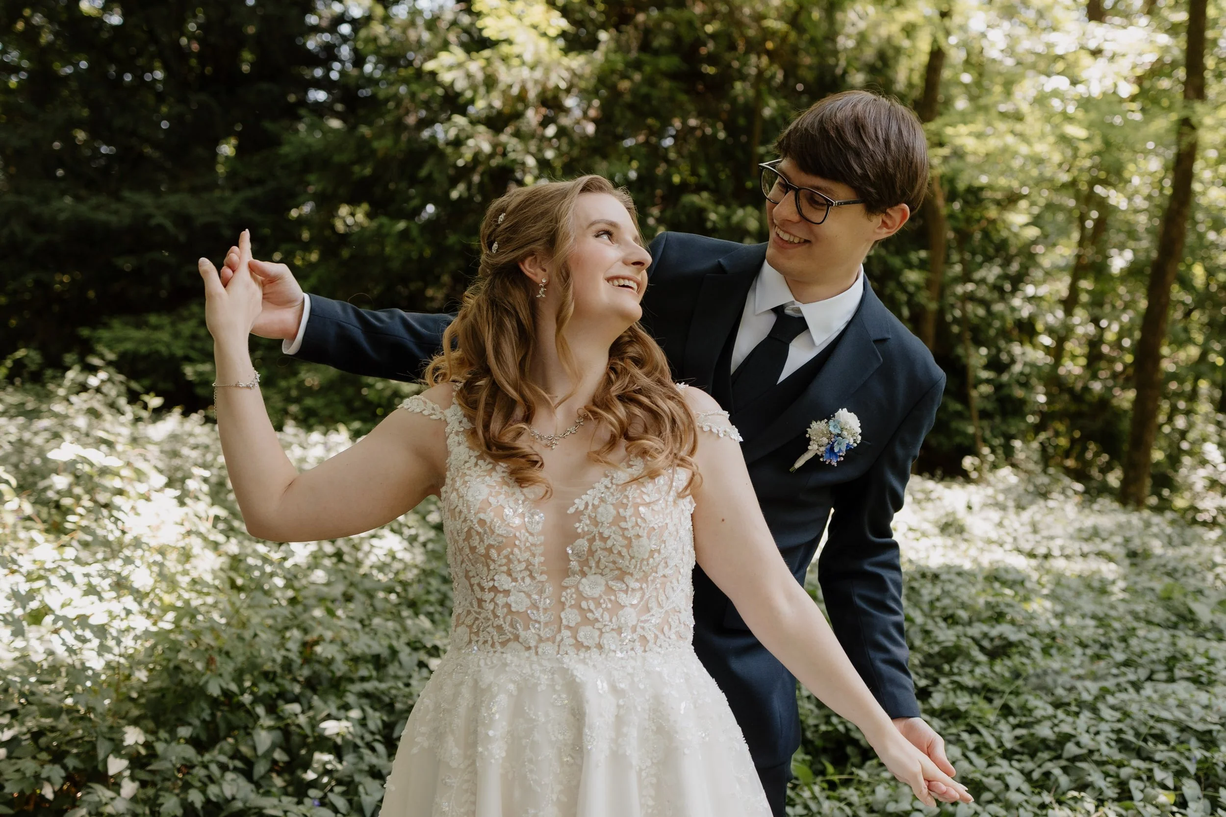 Ein Brautpaar tanzt im Wald, die Braut trägt ein weißes Hochzeitskleid mit Spitze, der Bräutigam trägt einen dunklen Anzug mit Krawatte. Sie lachen und schauen sich liebevoll an.