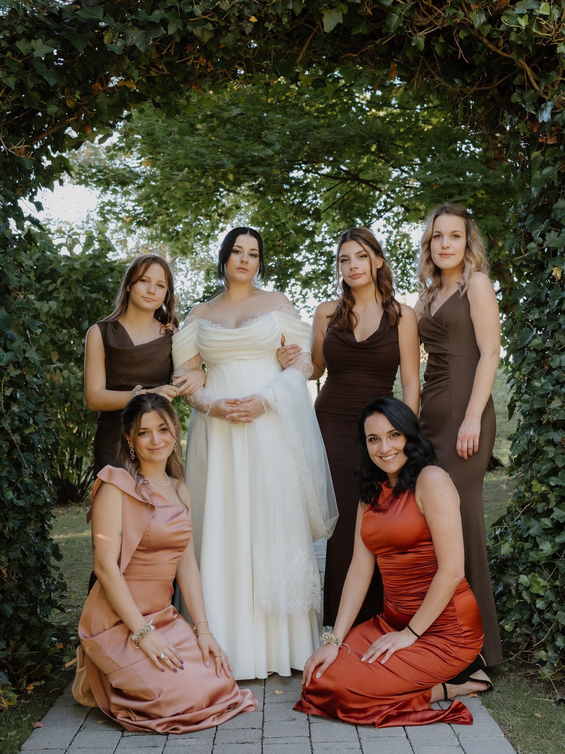 Eine Gruppe von Damen in eleganten Kleidern, einige sitzen, andere stehen, im Freien unter einem Baum bei einer Hochzeit.
