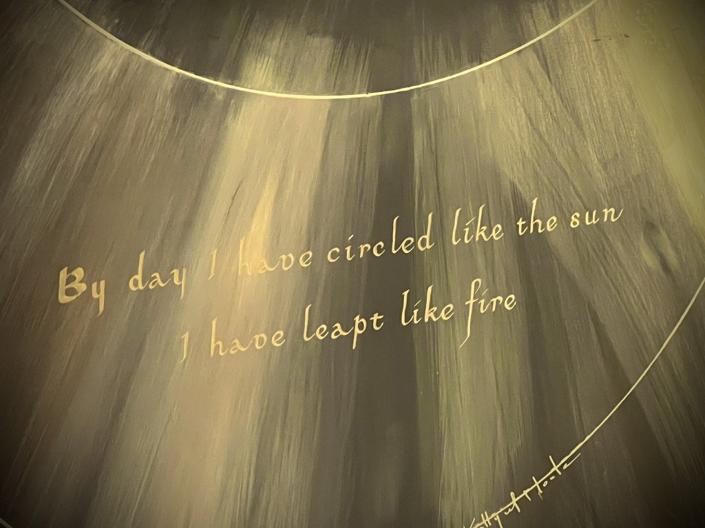 A mural with a quote written in gold script on a dark, abstract background with streaks of yellow and green. The quote says, 'By day I have circled like the sun I have leapt like fire'.