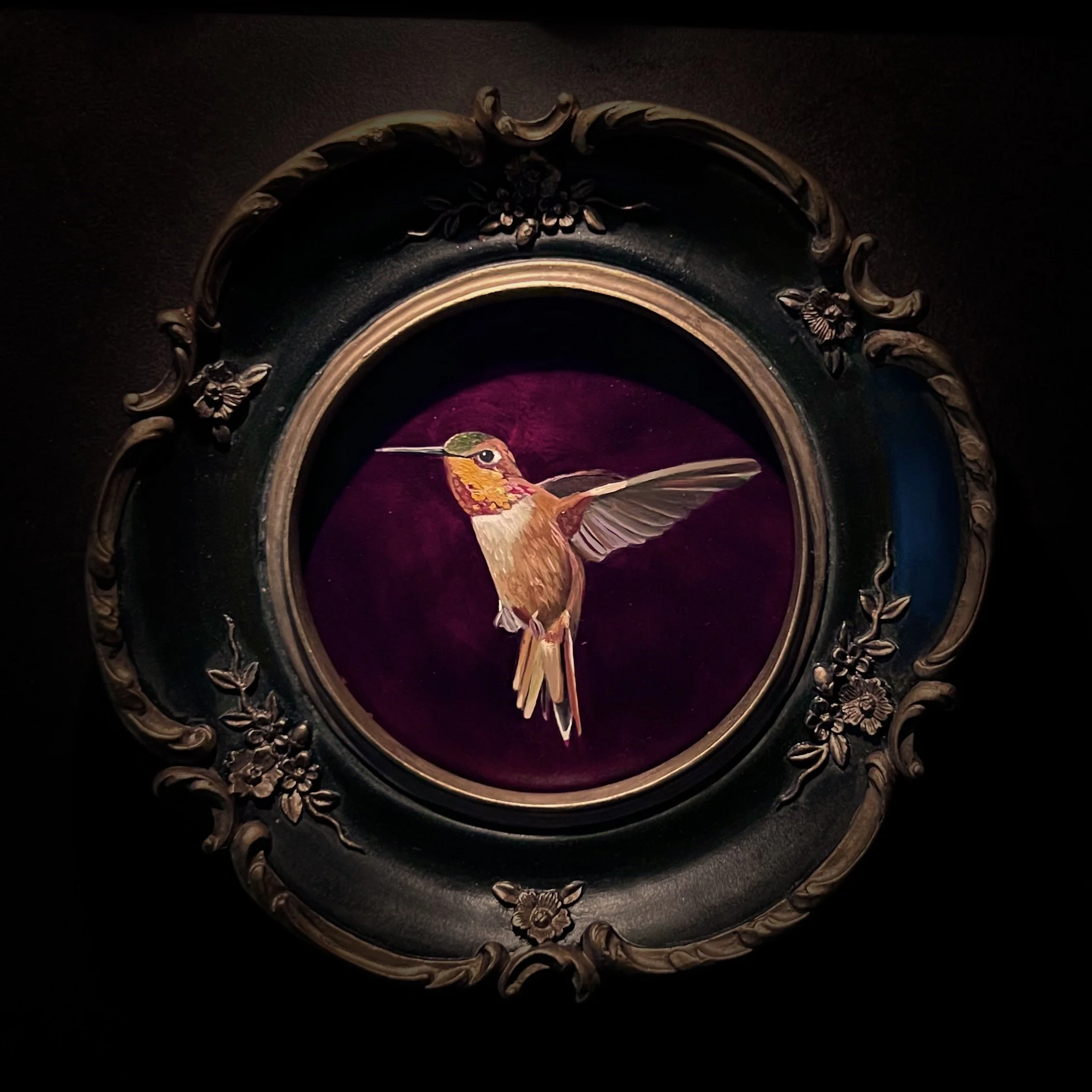 A painting of a colorful hummingbird on a decorative oval frame with floral details, against a dark background.