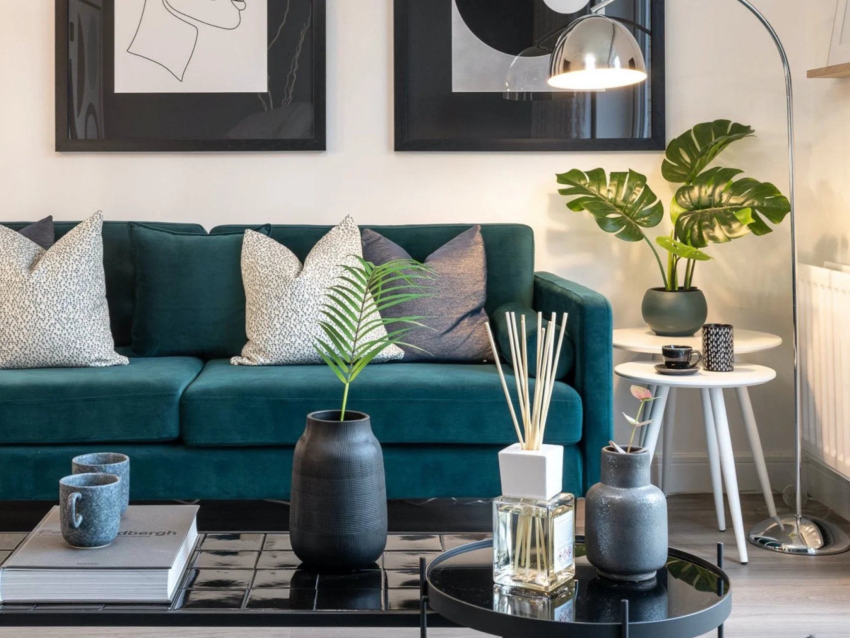 Living room with green sofa, decorative pillows, black framed wall art, potted plants, white side table with cups, and a black coffee table with vases and books.