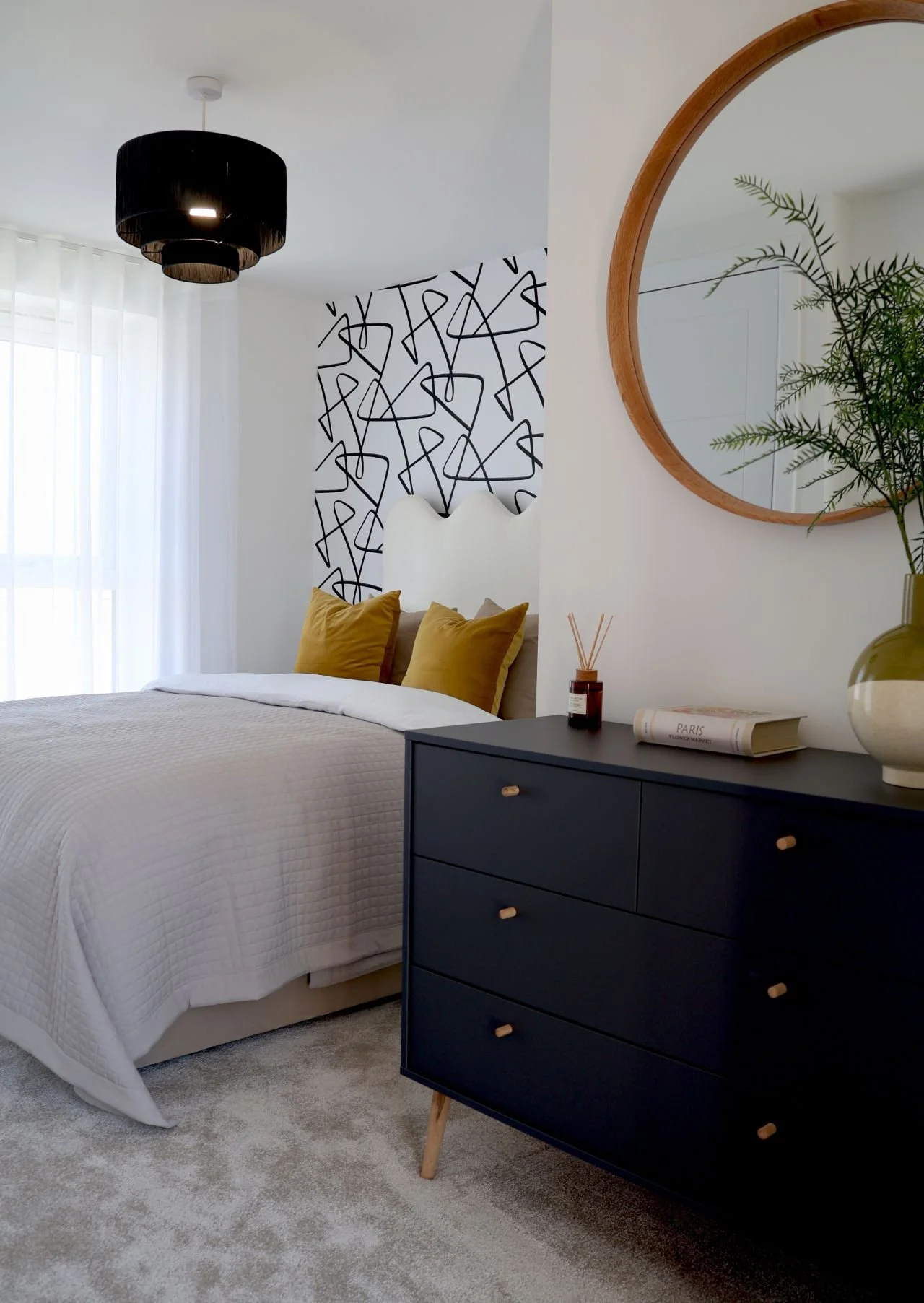 A modern bedroom with white walls, a bed with white bedding and mustard-colored pillows, a black dresser with light wood knobs, a round wooden mirror, a green plant in a white and green vase, and a decorative wall pattern behind the headboard. Natura