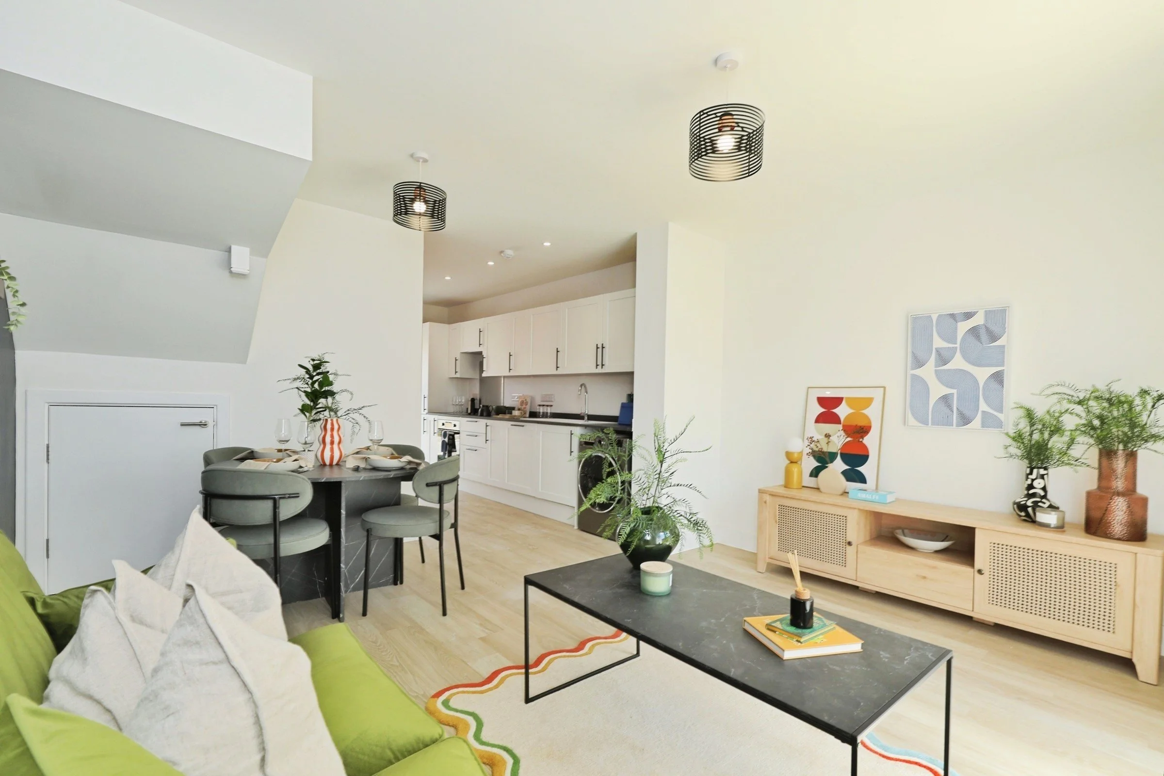 Modern living room with white walls, light wood flooring, a green couch, black coffee table, sideboard with decorative art and plant, and a kitchen in the background.