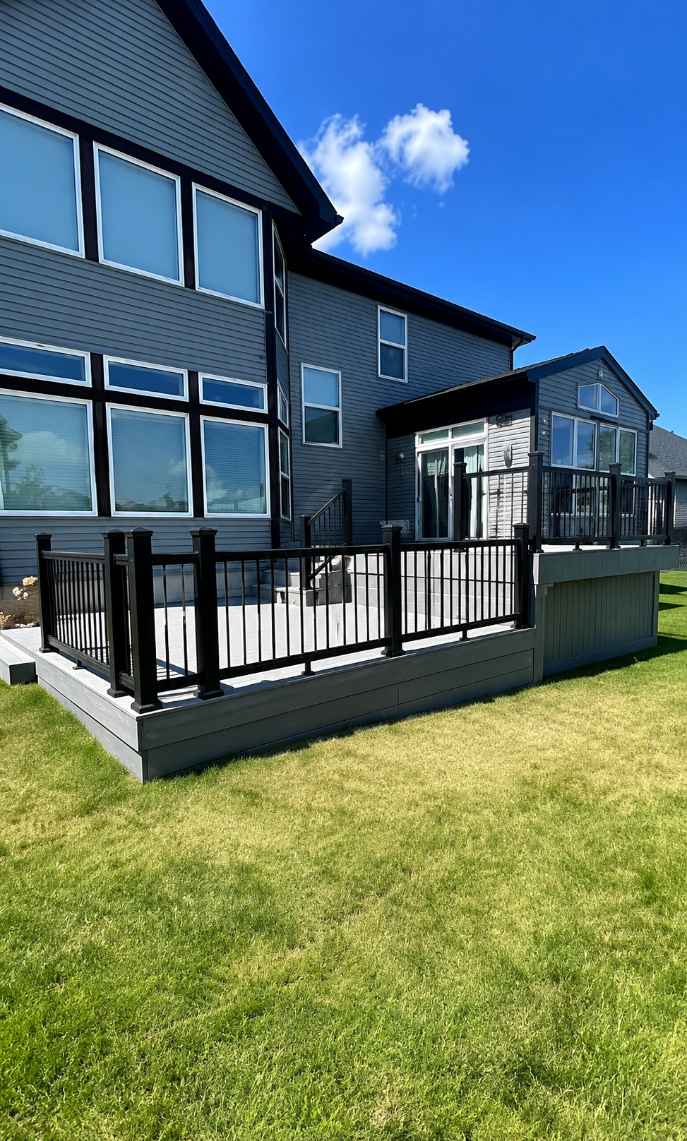 Backyard deck with black railing and sliding glass doors, attached to a gray house with multiple windows, under a clear blue sky.