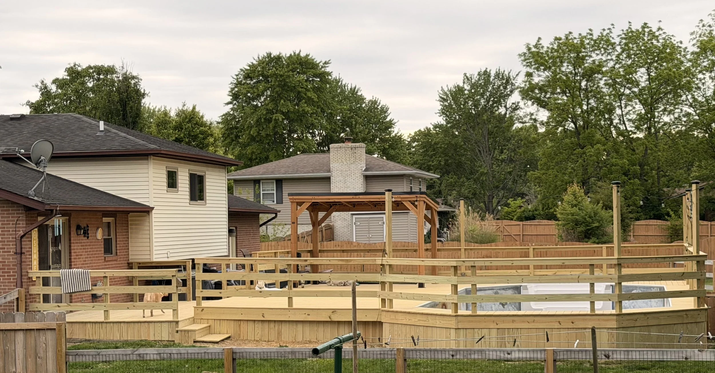 Residential backyard with a wooden deck under construction, neighboring houses, a wooden fence, lush green trees, and a cloudy sky.