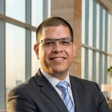 A young man in a suit and tie, wearing glasses, smiling in an office setting with large windows in the background.